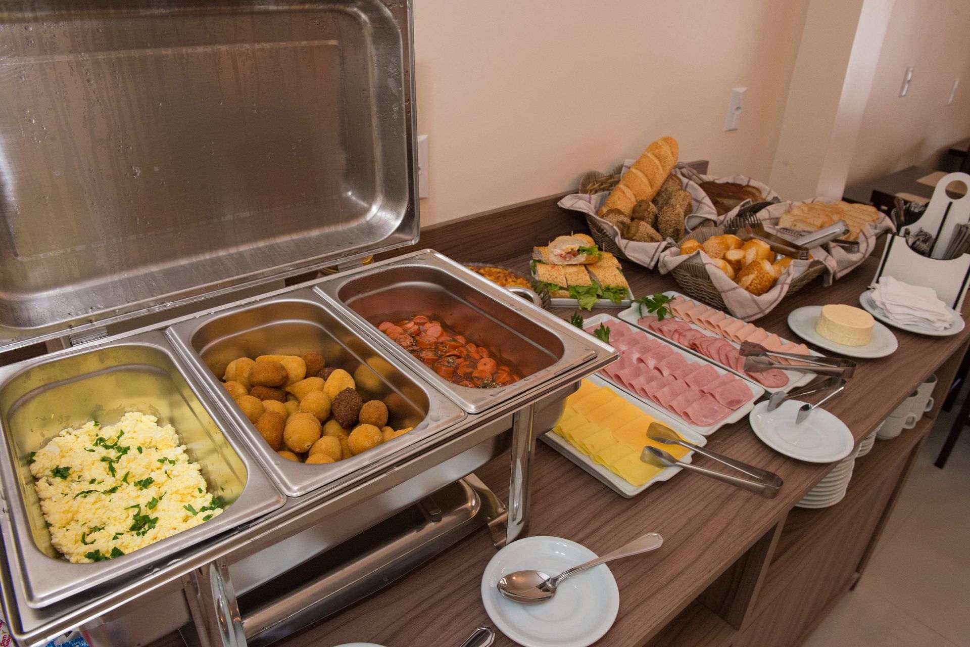 Buffet de café da manhã com ovos mexidos, batatas, feijão, pães, carnes e queijos.