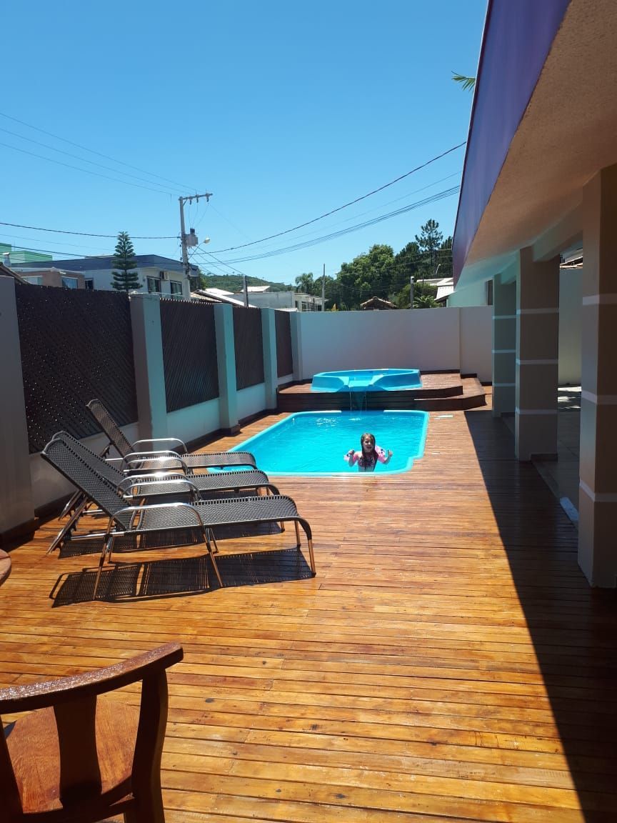 Área da piscina: pessoa na piscina azul, deck de madeira com espreguiçadeiras, spa azul, cerca e prédio sob um céu ensolarado.