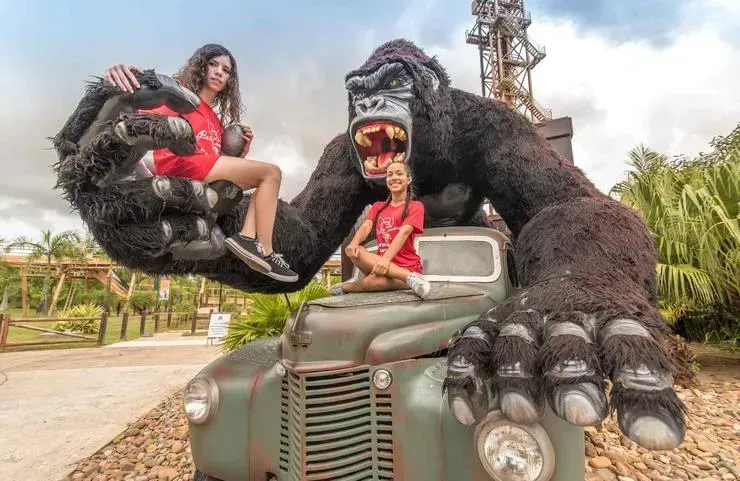 Duas jovens sentadas em um caminhão de brinquedo, seguradas por uma grande figura do King Kong. Elas vestem camisetas vermelhas e sorriem do lado de fora, com árvores e uma torre de metal ao fundo.