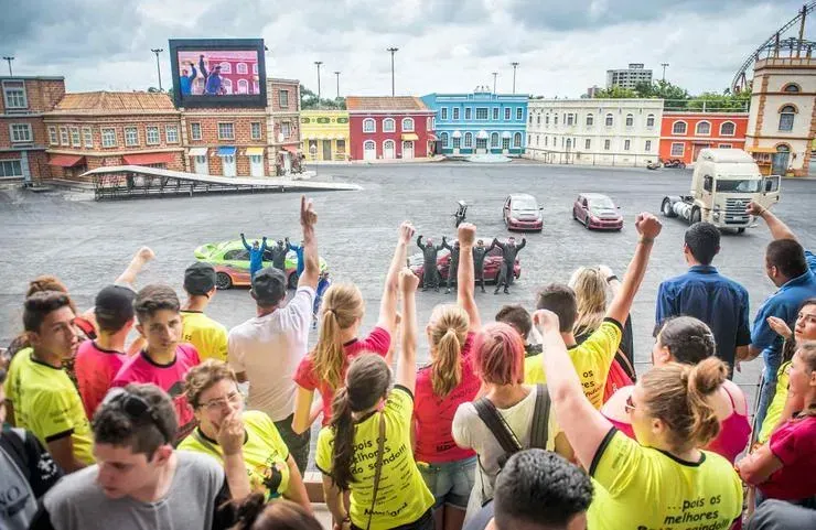Uma multidão comemora em frente a uma cidade simulada e colorida com veículos. Muitas pessoas levantam os braços, algumas vestindo camisetas amarelas.