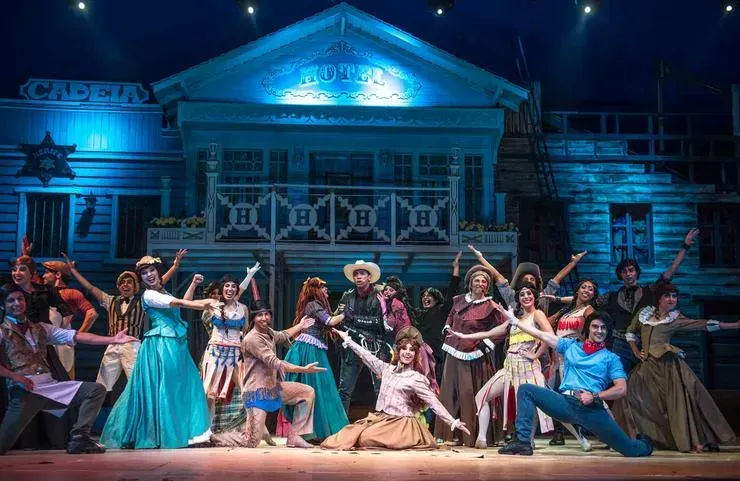 Elenco de teatro musical em trajes de faroeste no palco, em frente a um cenário de salão, realizando uma dança.
