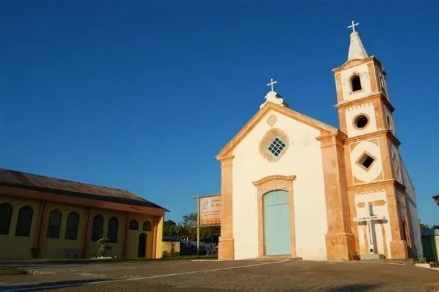 Igreja com torre sineira contra um céu azul brilhante. O edifício é creme e laranja, com uma cruz na fachada.