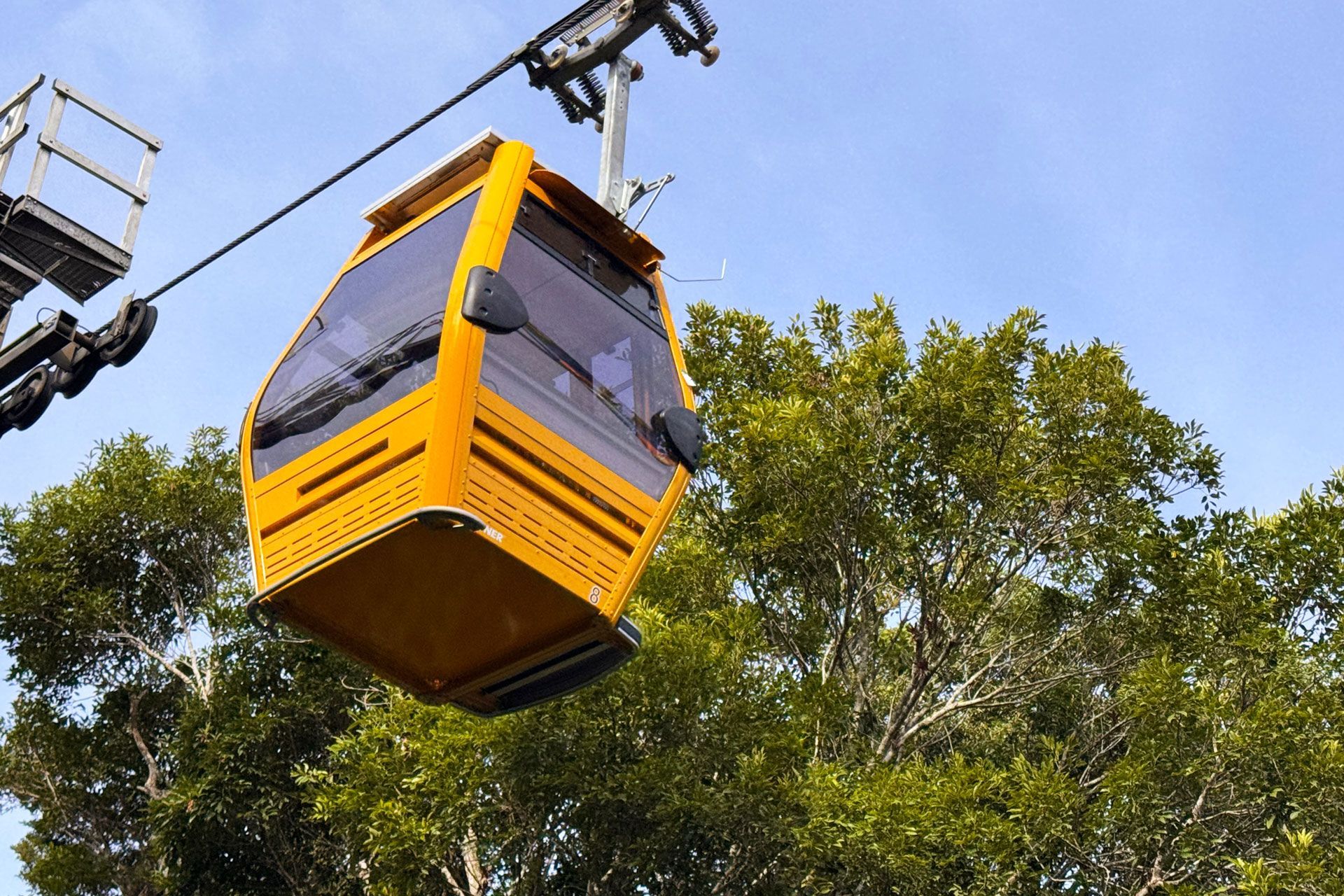 Um teleférico amarelo suspenso por um cabo contra um céu azul. O teleférico passa sobre árvores verdes.