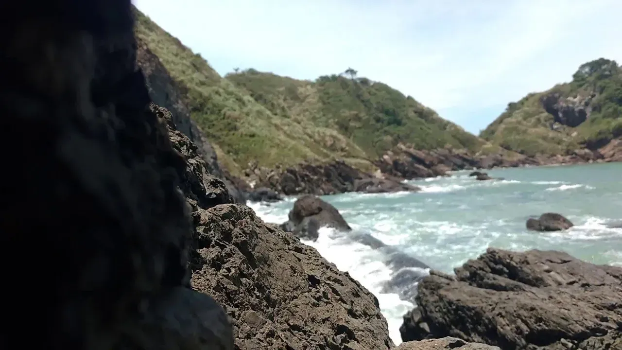 Litoral rochoso com ondas quebrando, colinas verdes ao fundo e um céu azul.