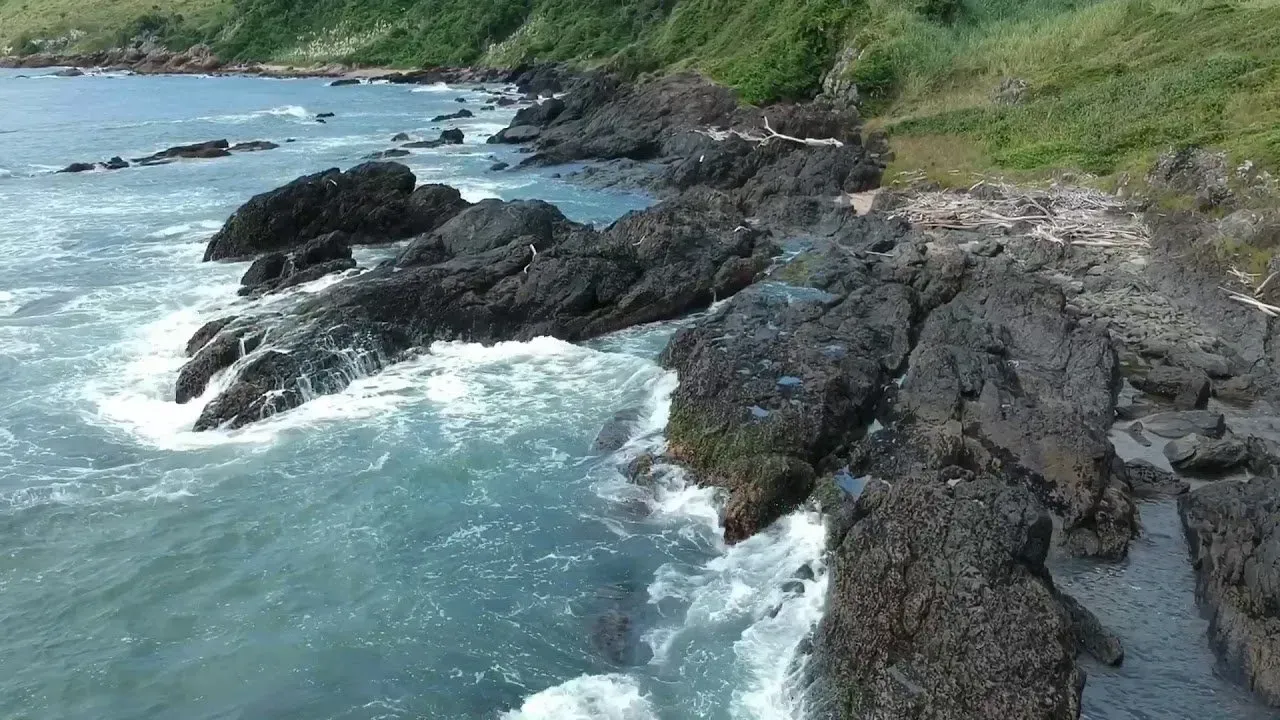 Litoral rochoso com ondas quebrando contra rochas escuras e vegetação verde ao fundo.