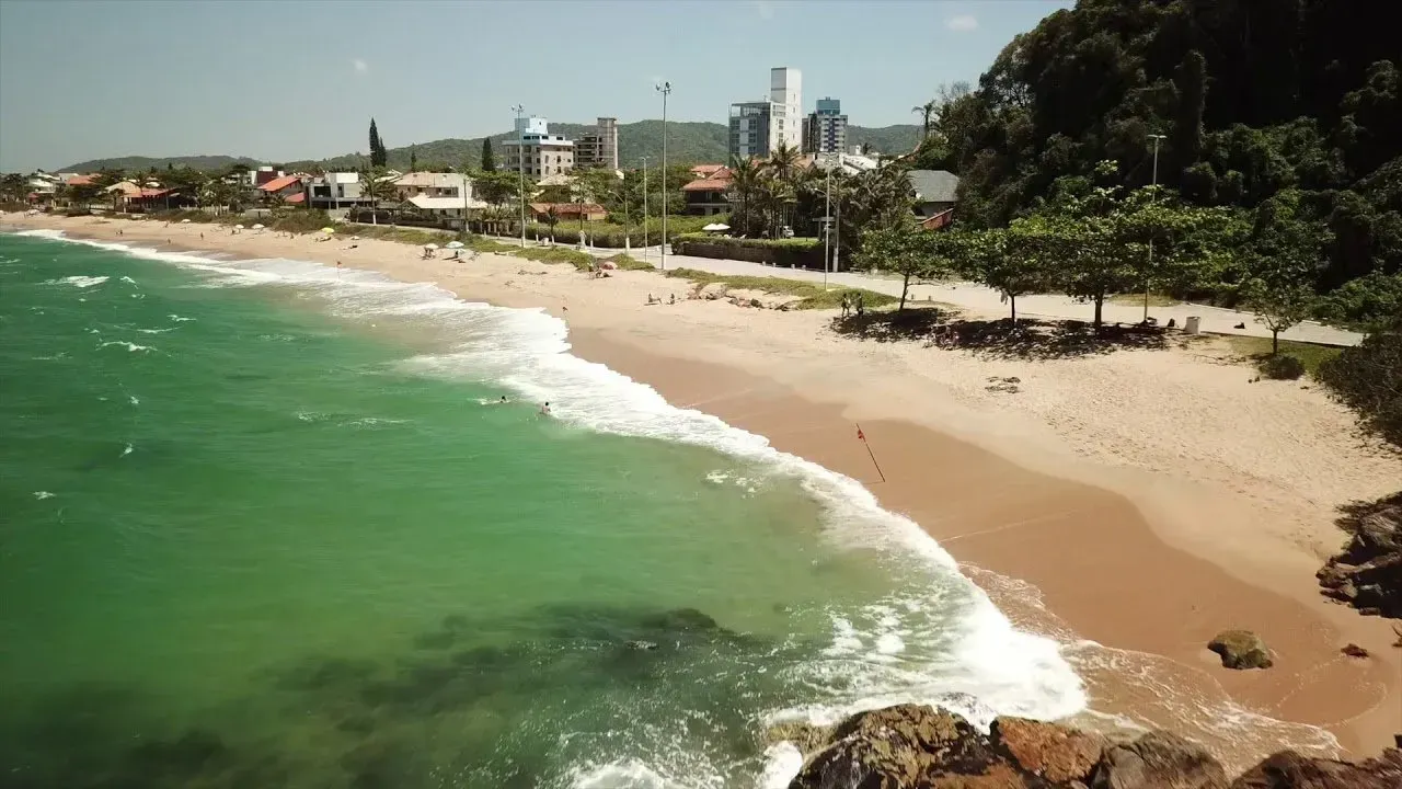Praia com água azul-turquesa, areia dourada e um horizonte de edifícios ao fundo em um dia ensolarado.