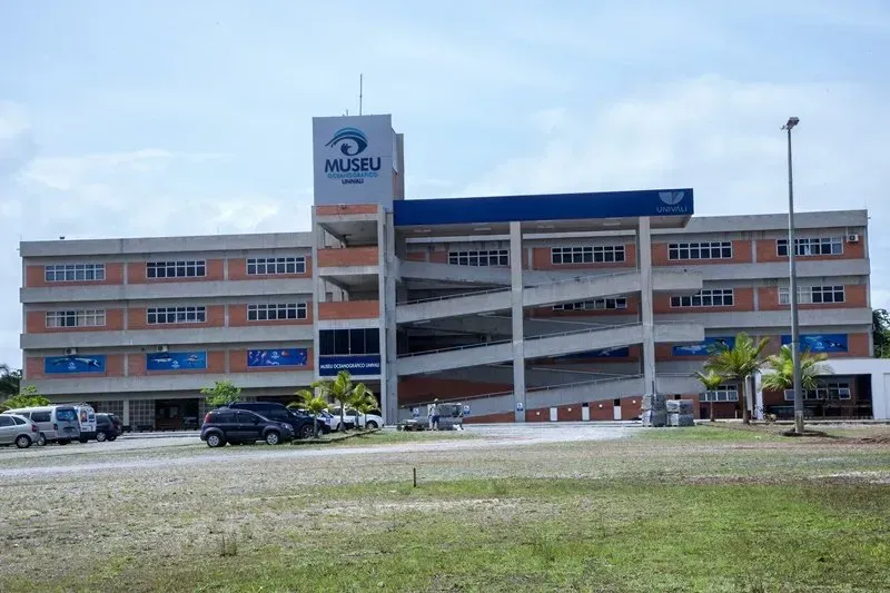 Prédio de três andares com rampa, logotipo da MUDBU e estacionamento. Carros cinzas estacionados em frente ao prédio.