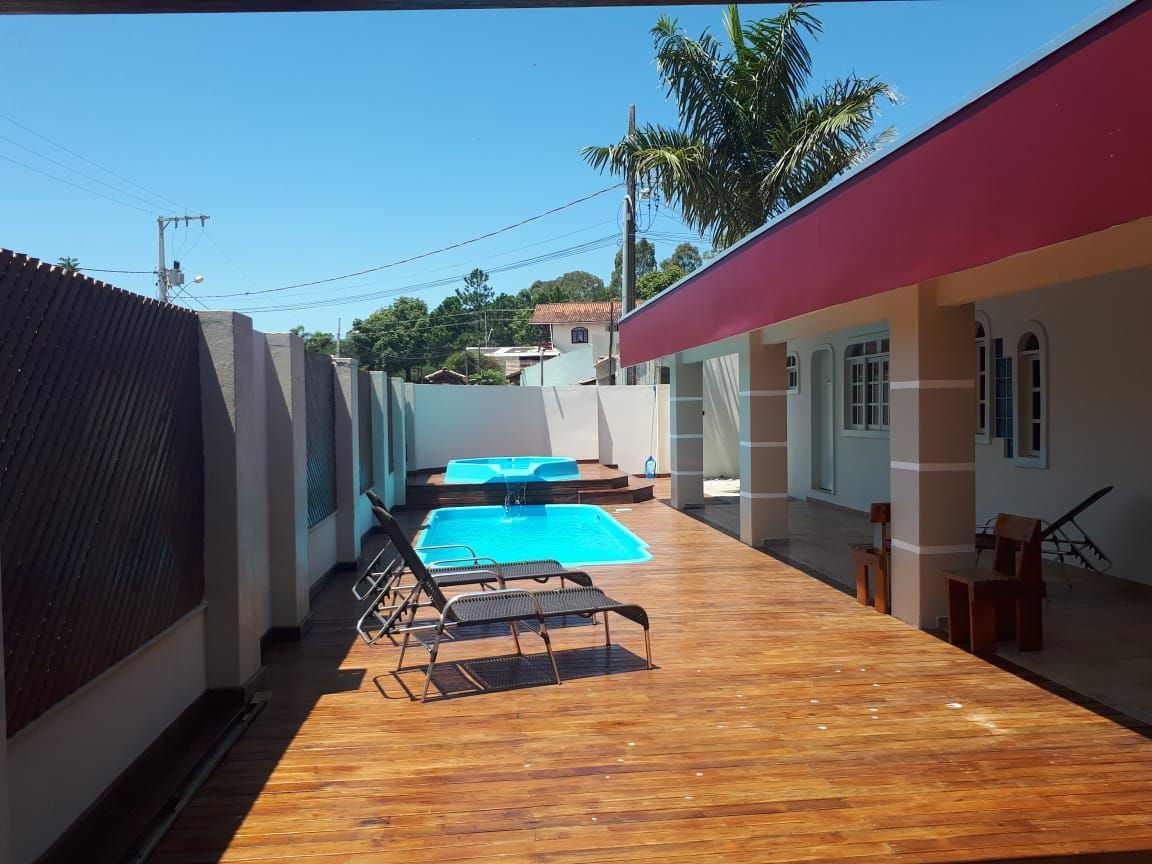 Um quintal com deck de madeira, duas piscinas e uma casa com telhado vermelho e colunas brancas. Espreguiçadeiras ficam perto da piscina.
