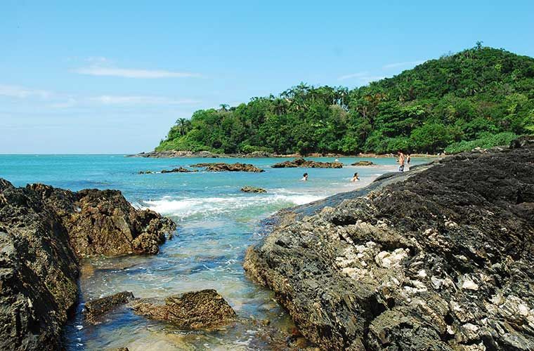 Litoral rochoso com águas azul-turquesa, praia de areia e uma colina verdejante sob um céu azul.