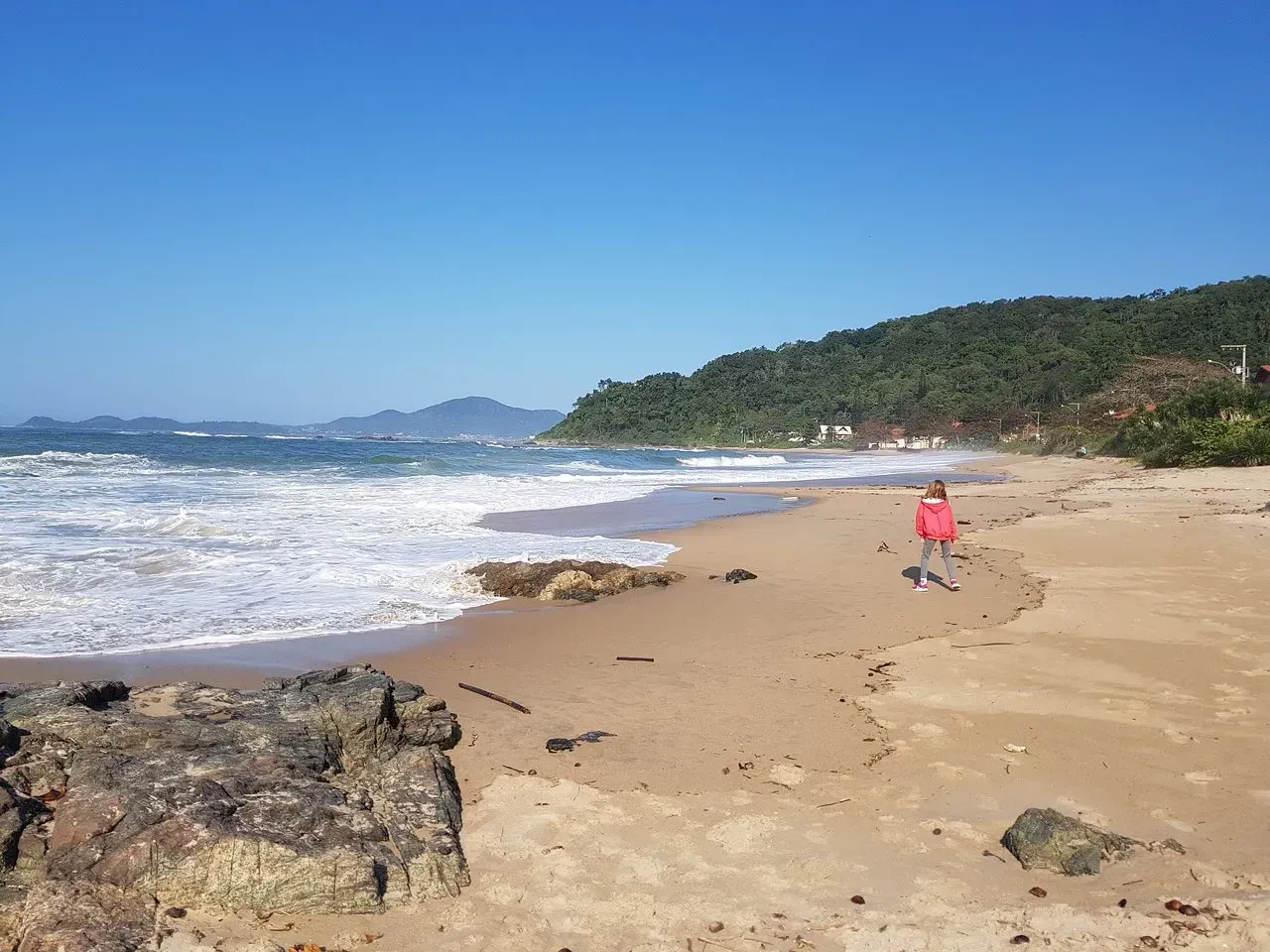 Uma cena de praia ensolarada com ondas quebrando na praia. Uma pessoa de vermelho caminha pela areia e, ao fundo, uma encosta arborizada.