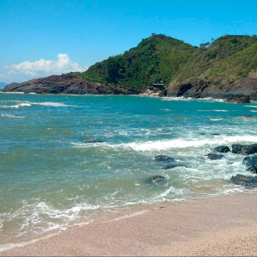 Praia de areia com águas azul-turquesa e um cenário verdejante e montanhoso sob um céu azul claro.