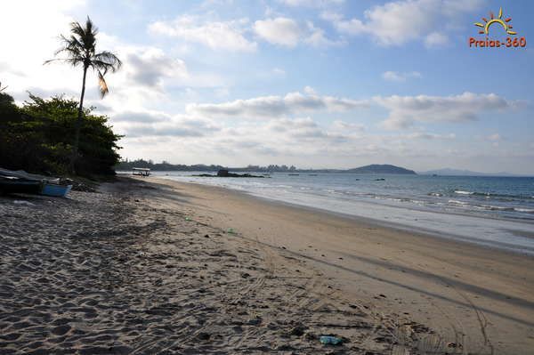 Praia de areia com palmeiras e barcos no horizonte sob um céu parcialmente nublado.