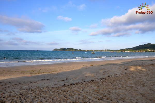 Praia de areia com água azul, uma pequena ilha ao longe e um céu parcialmente nublado.