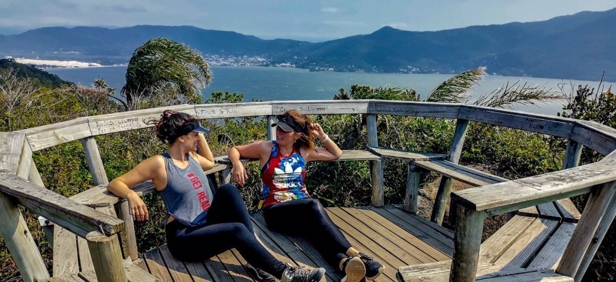 Duas mulheres estão sentadas em um banco de madeira com vista para um corpo d'água.