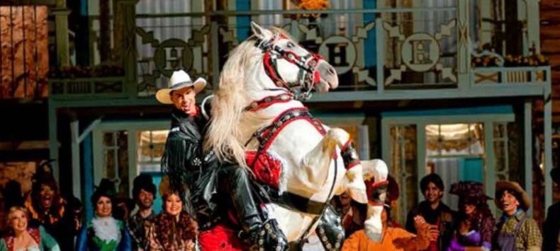 Um homem com chapéu de cowboy está montando um cavalo branco na frente de uma multidão.