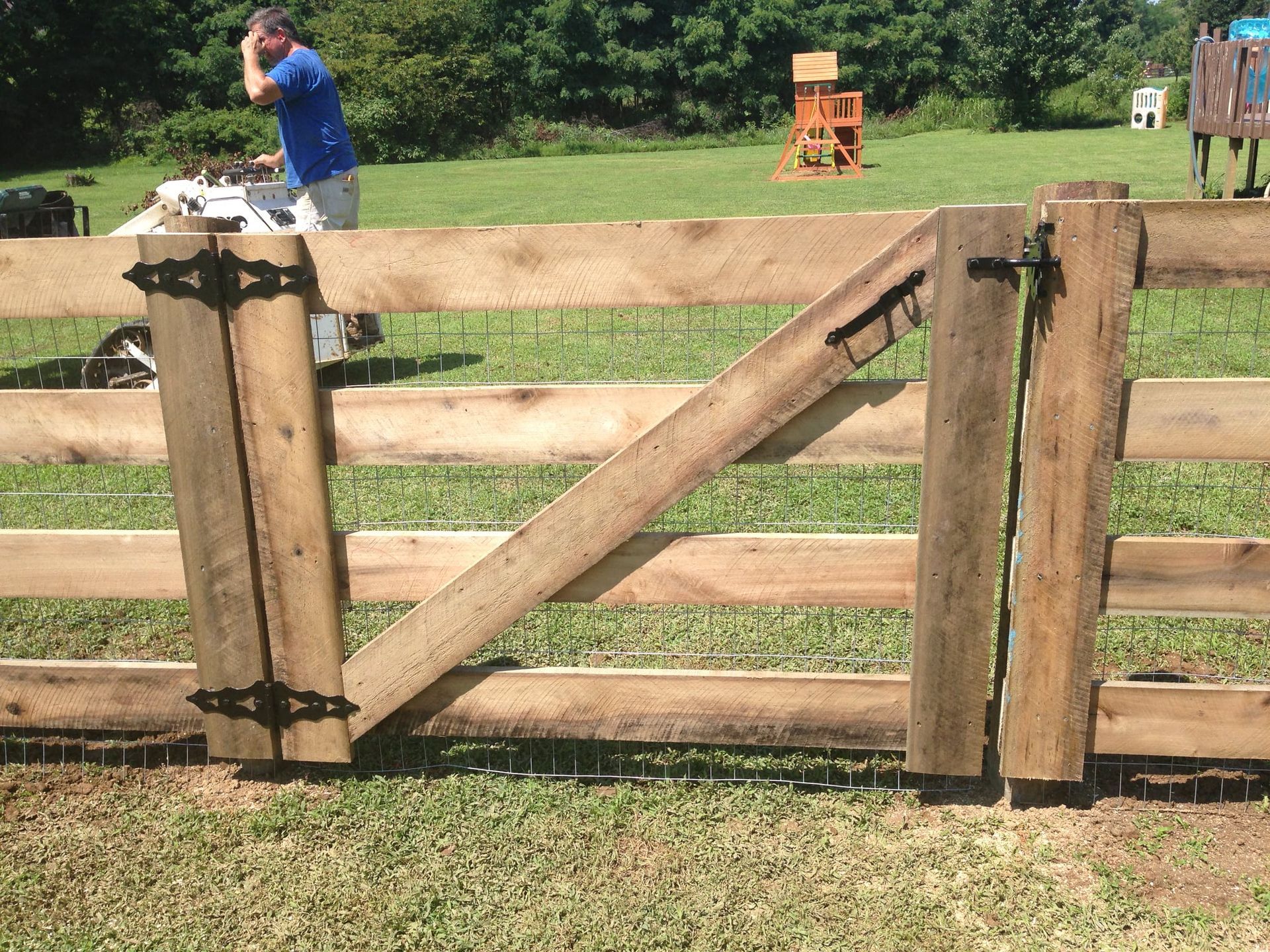 Wooden farm gate with diagonal brace in a grassy field; a person stands in the background.