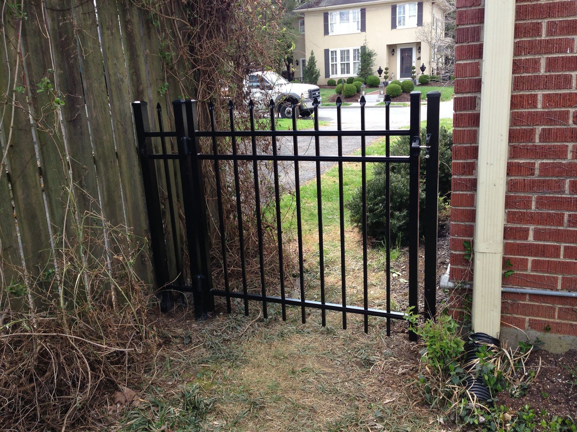 Black metal gate in a narrow overgrown path beside a red brick house and wooden fence.