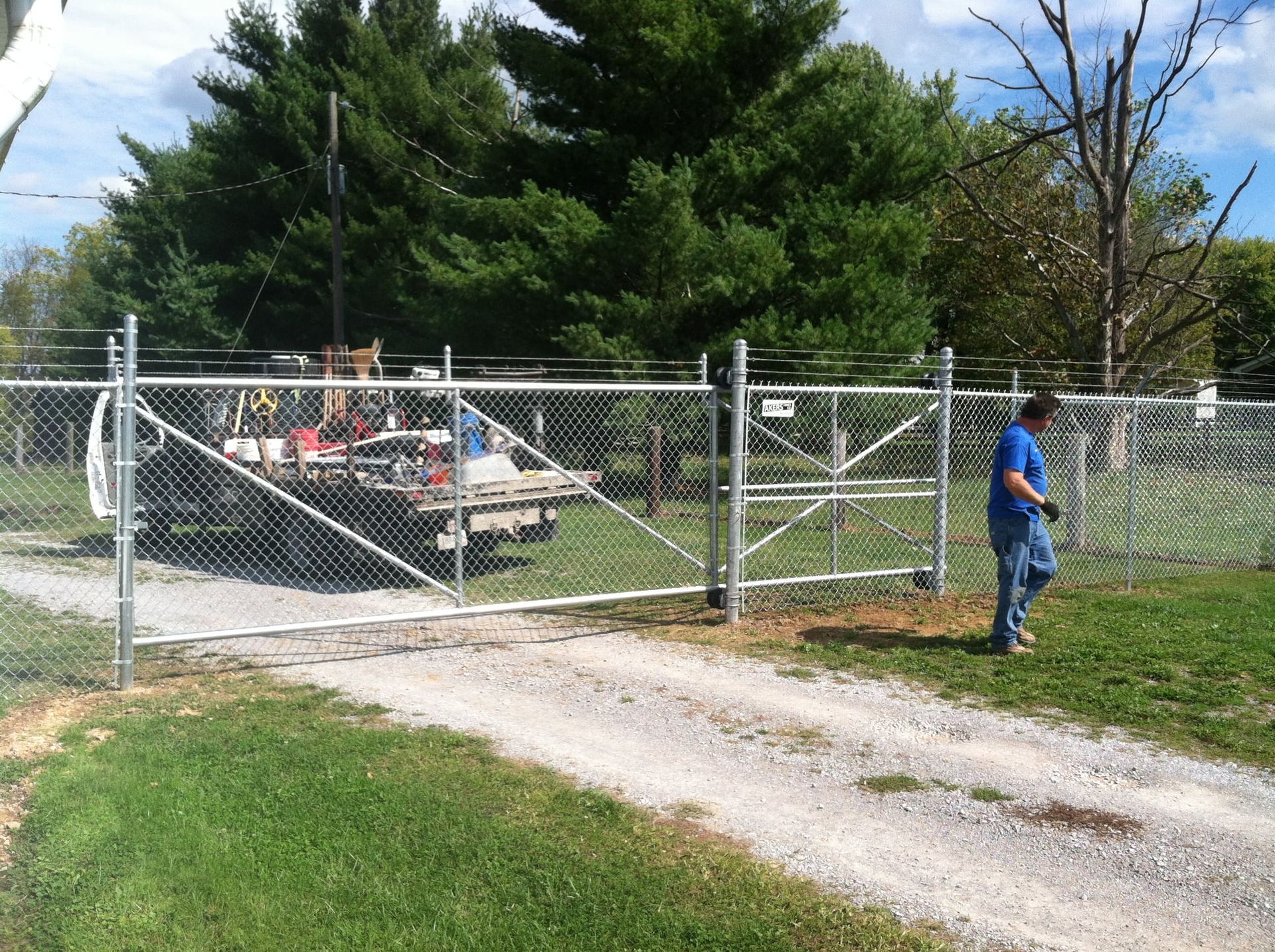 Chain-link gate blocking a gravel driveway, with a person standing beside it near trees and a utility trailer.