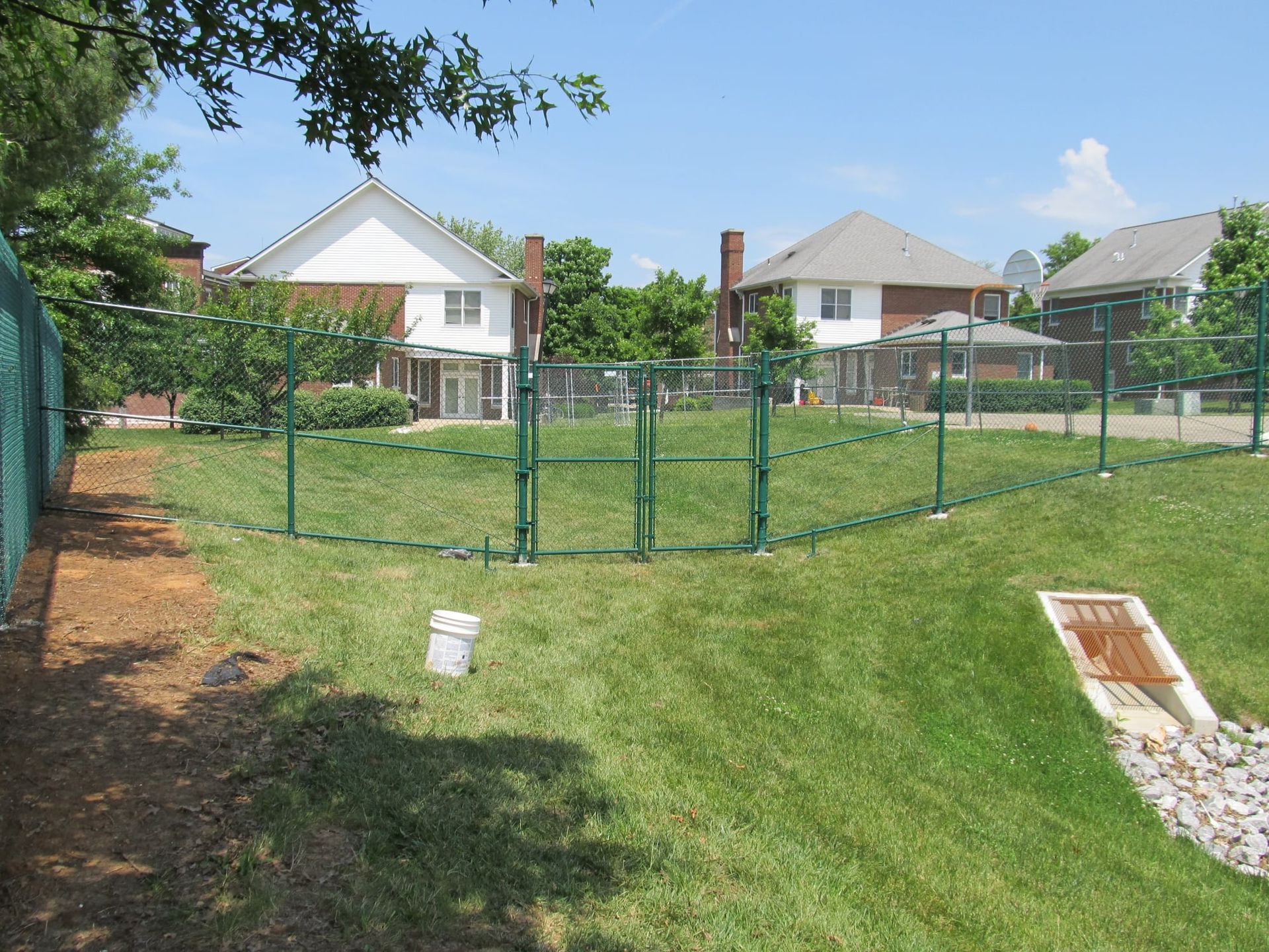 Backyard with green metal fence, grassy hill, and suburban houses under a clear blue sky