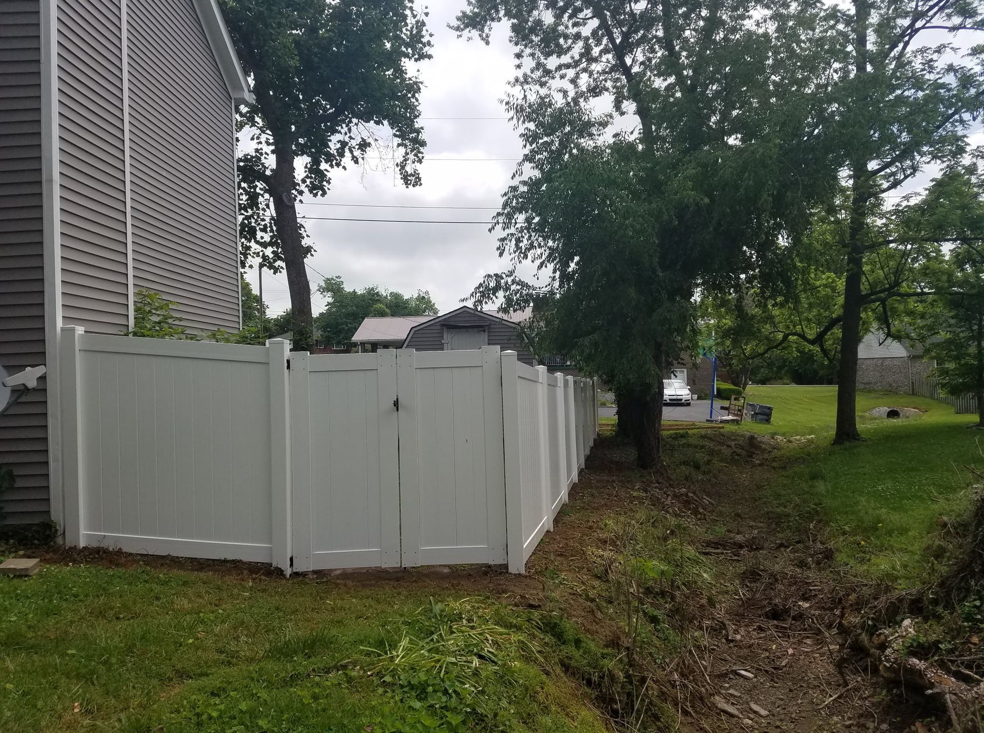 White vinyl fence beside a house, with a muddy drainage ditch and trees in a grassy yard.