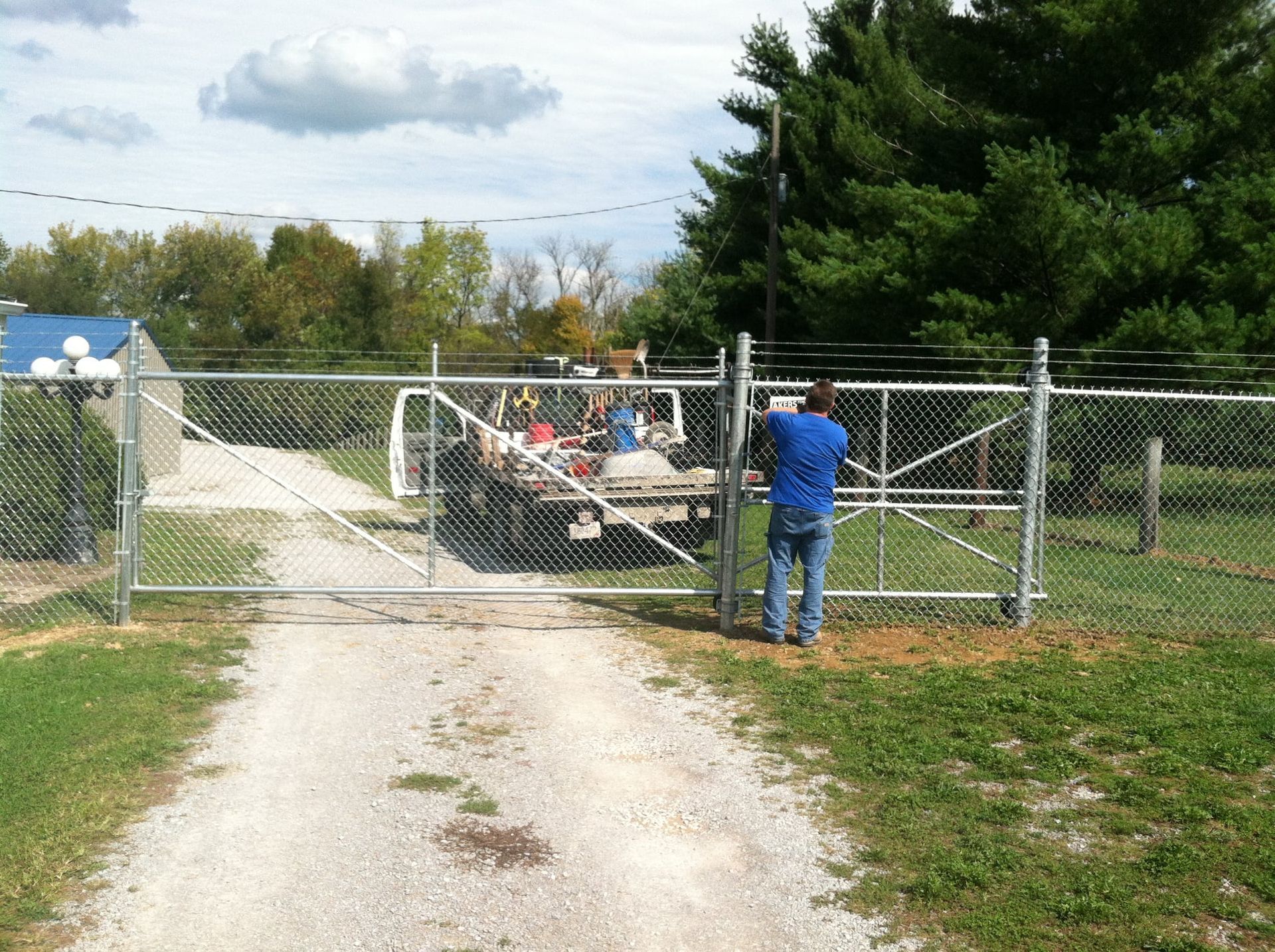 People standing by a chain-link gate at a gravel driveway, with a vehicle and trees behind it