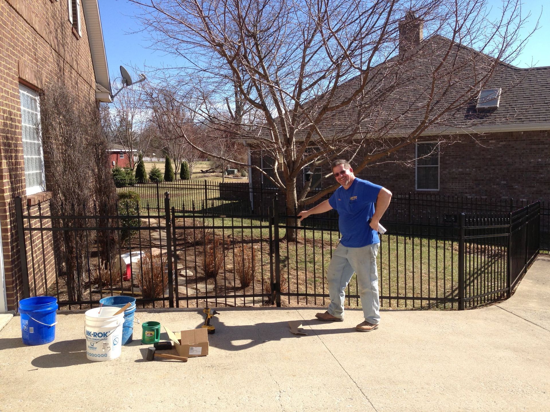 Person standing in a driveway beside tools and buckets near a black metal fence and brick houses.