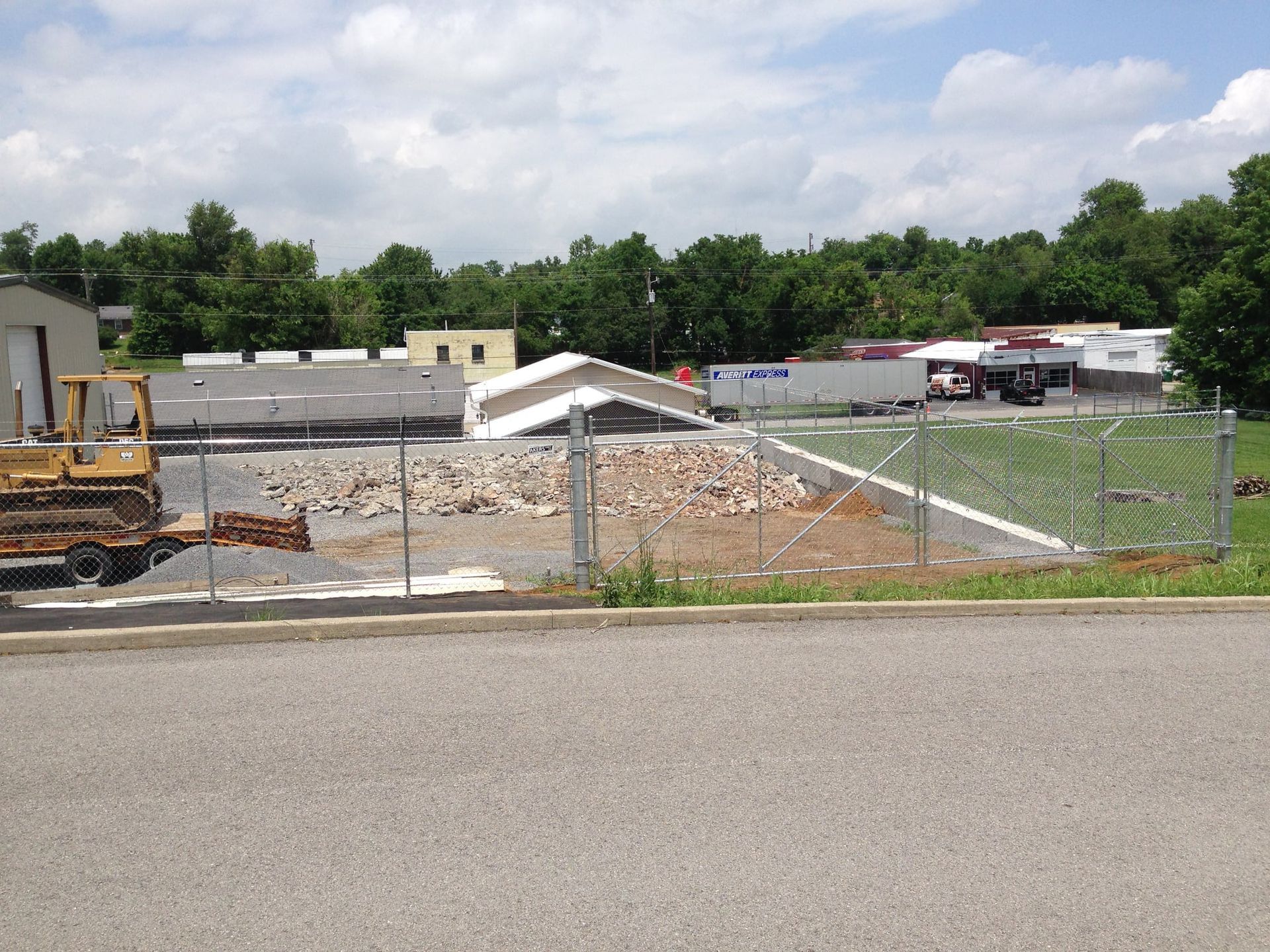 Construction site with excavator, rubble, and fenced-off area in front of low buildings and trees