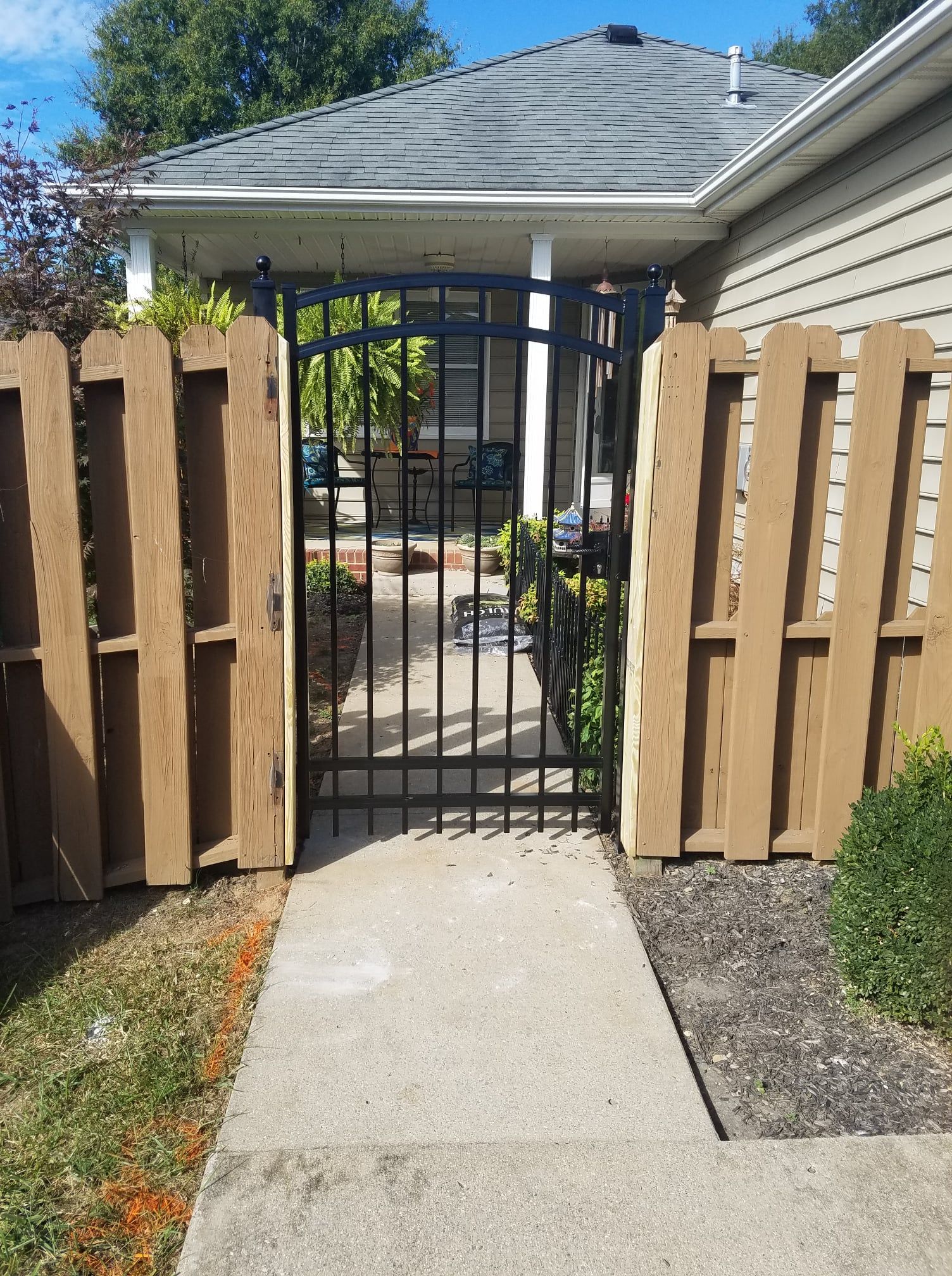 Black metal gate between wooden fence panels, opening onto a front walkway and house entrance.