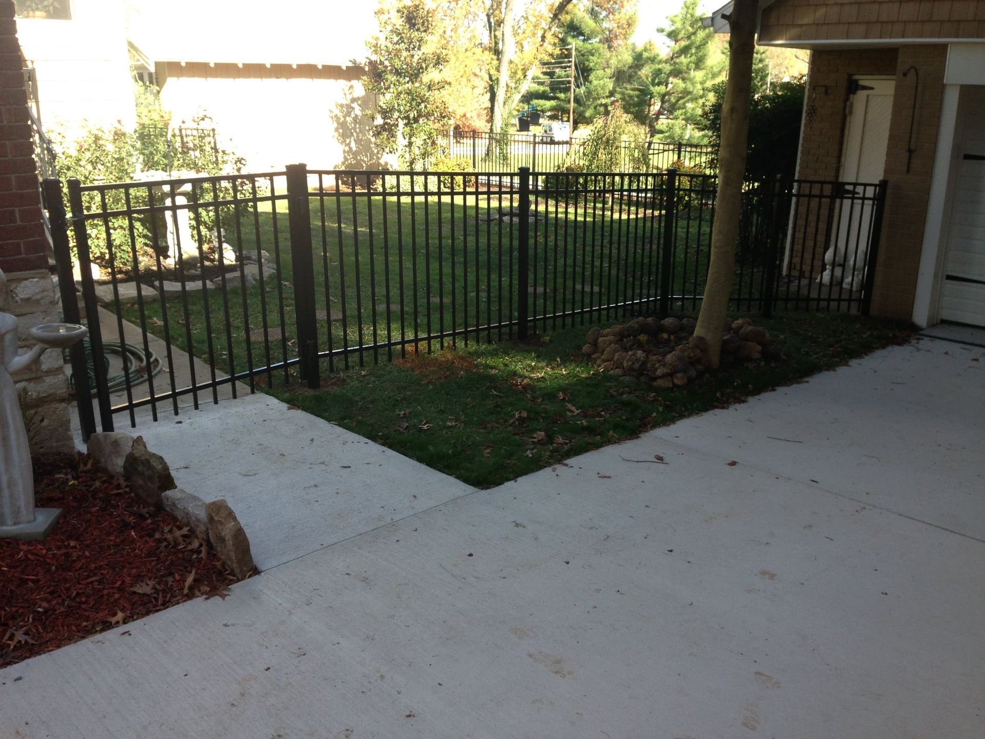 Apartment patio walkway beside a black metal fence and grassy courtyard, with concrete paths and trees.