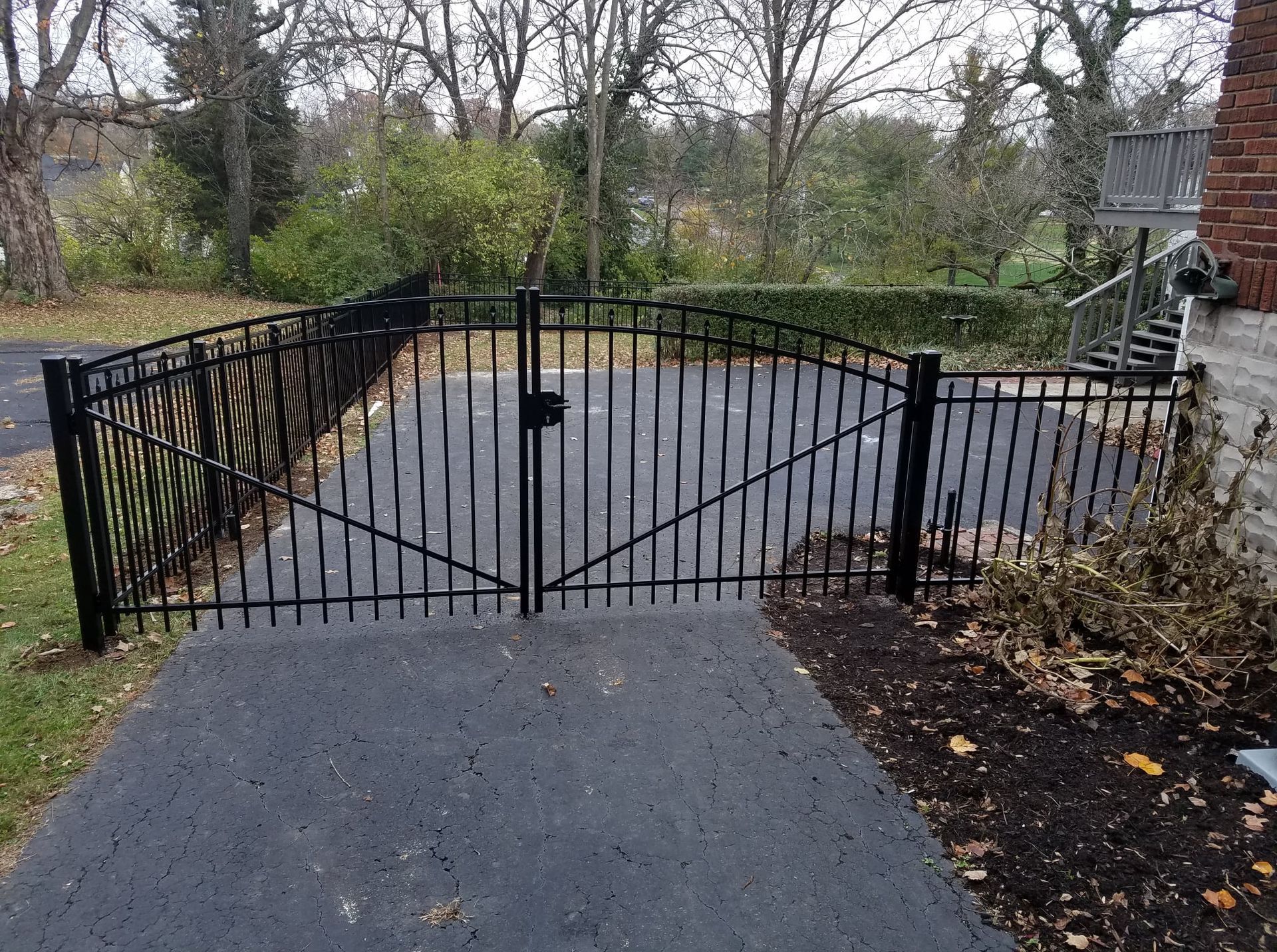 Black metal gate blocking a driveway, with trees and a brick house in the background