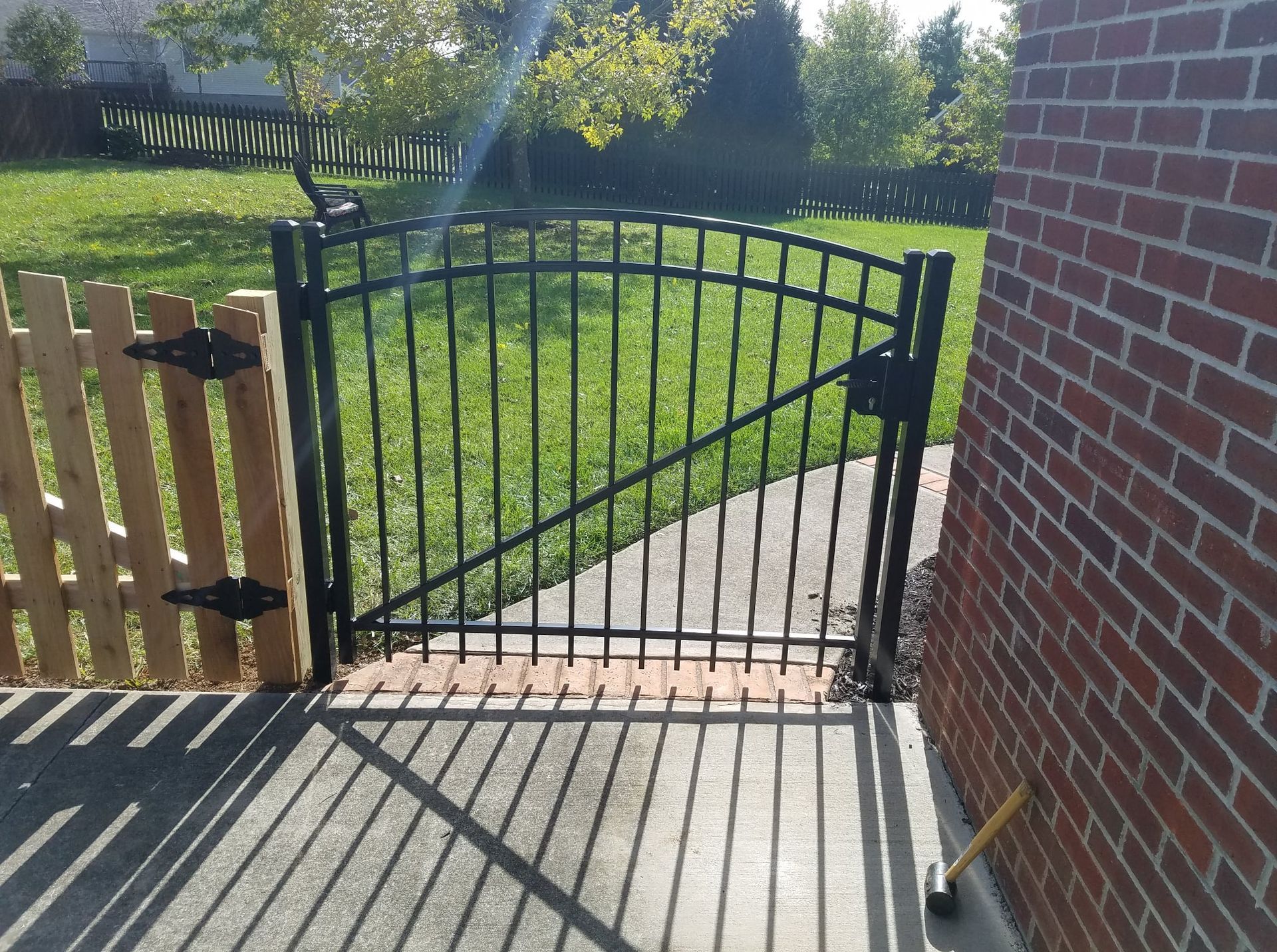 Black metal gate between a wooden fence and brick wall, opening onto a sunny grass lawn and walkway.