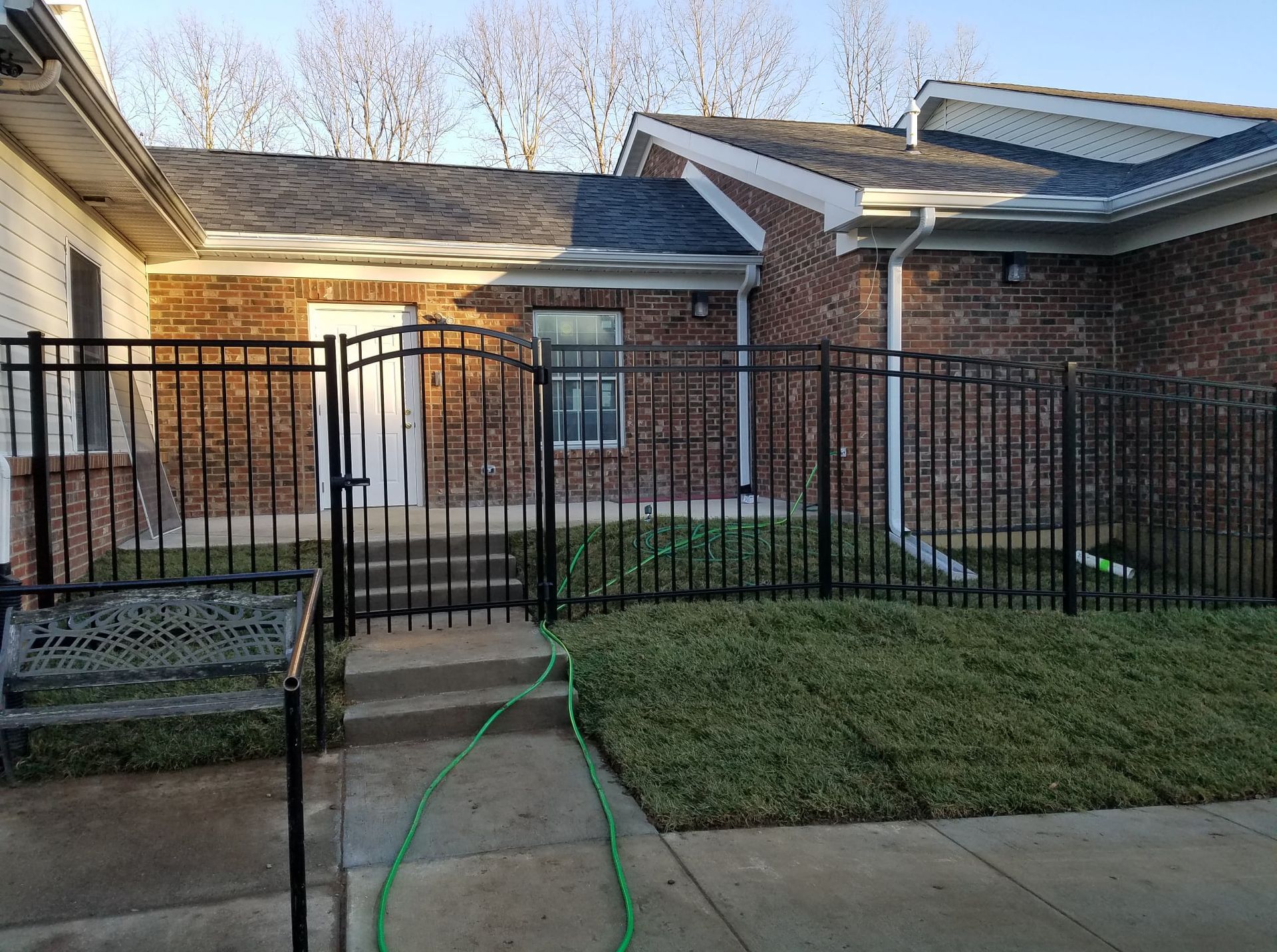 Brick house front yard with black metal fence, concrete walkway, and green hose on the grass