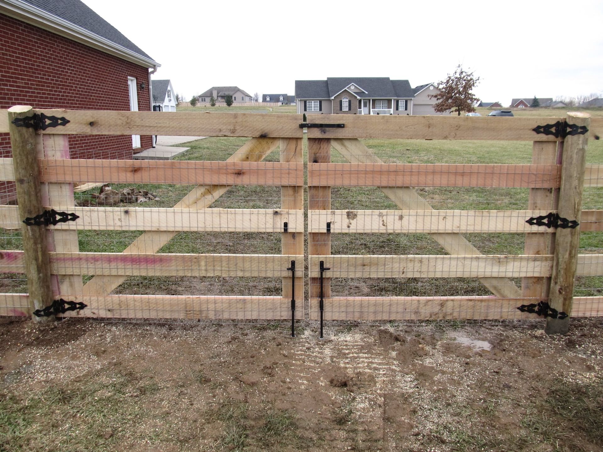 Wooden farm gate with wire mesh across a dirt path, backed by red-brick houses and overcast sky.