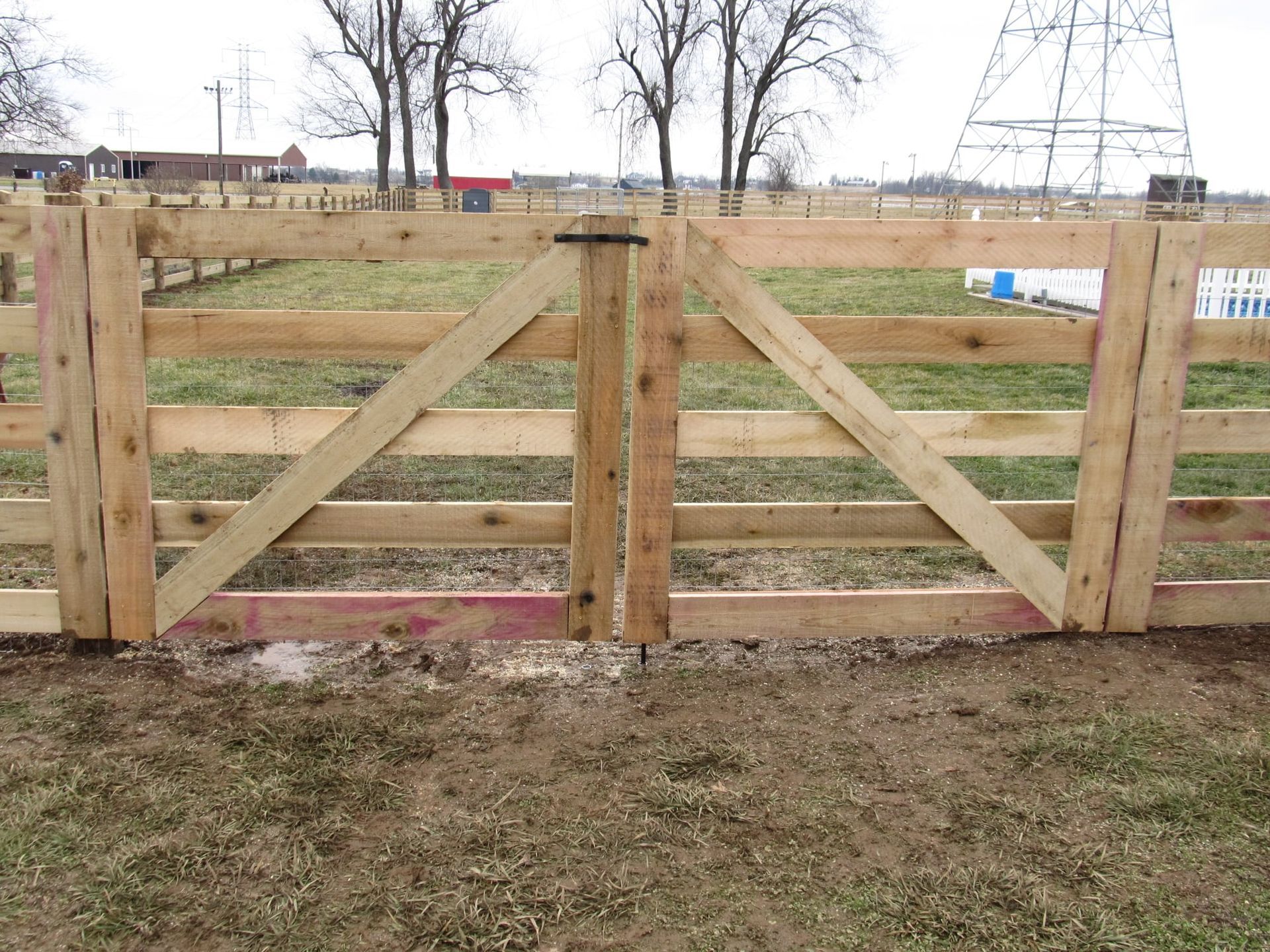Wooden farm gate with diagonal bracing in a muddy field, with bare trees and buildings in the background