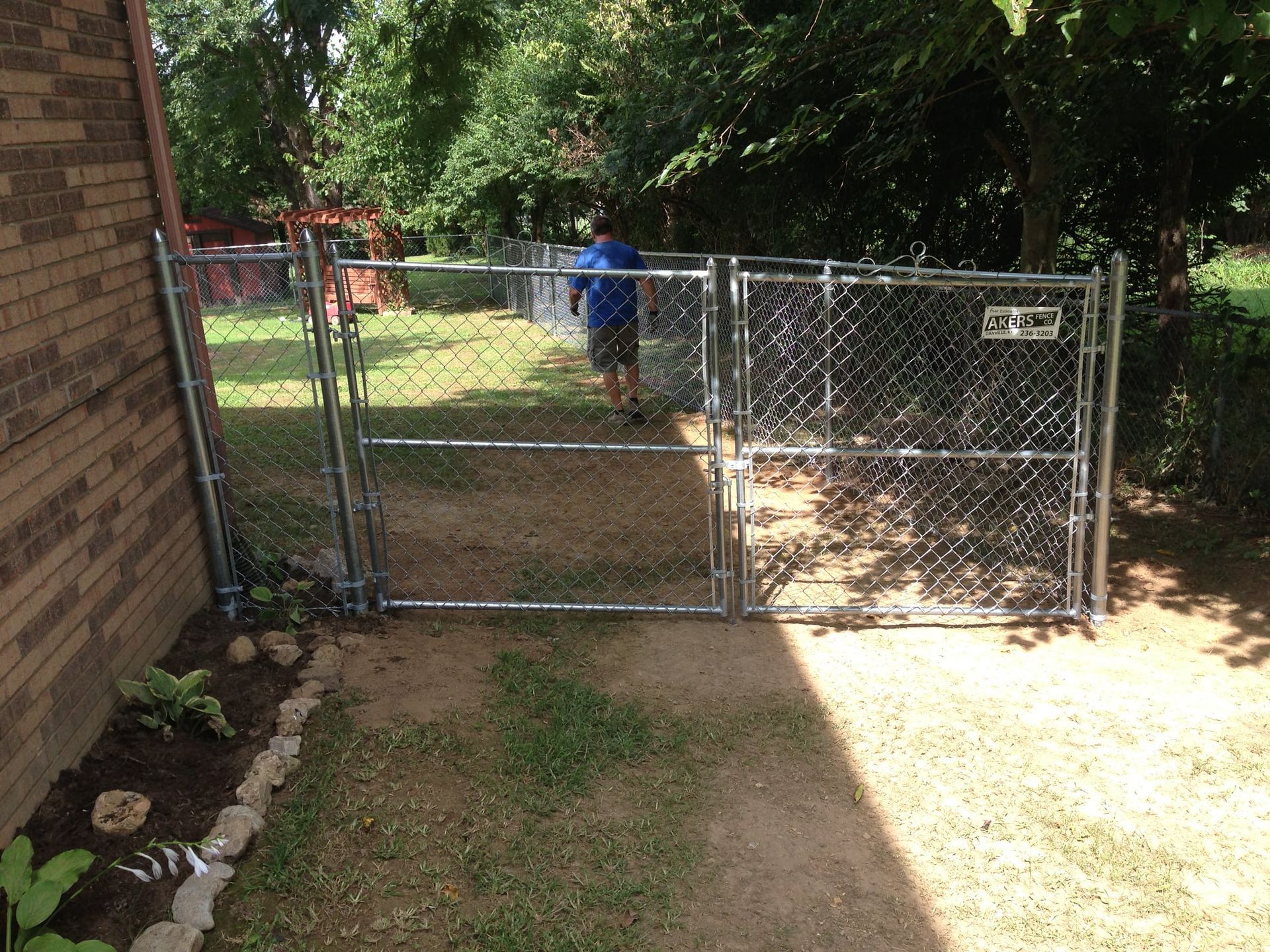 Chain-link gate beside a brick wall opening into a sunny yard with trees and a person in blue in the background