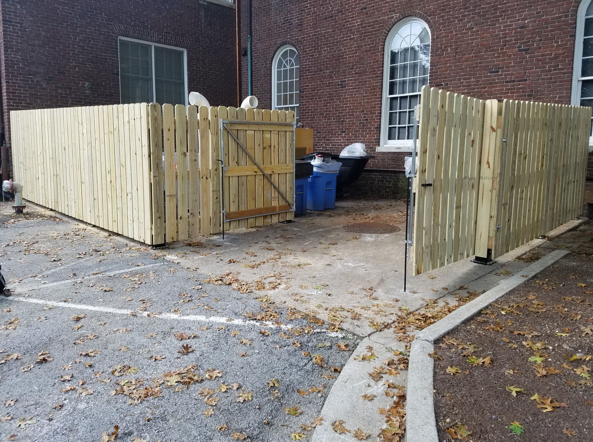 Wooden fence enclosure around dumpsters beside a brick building and parking lot