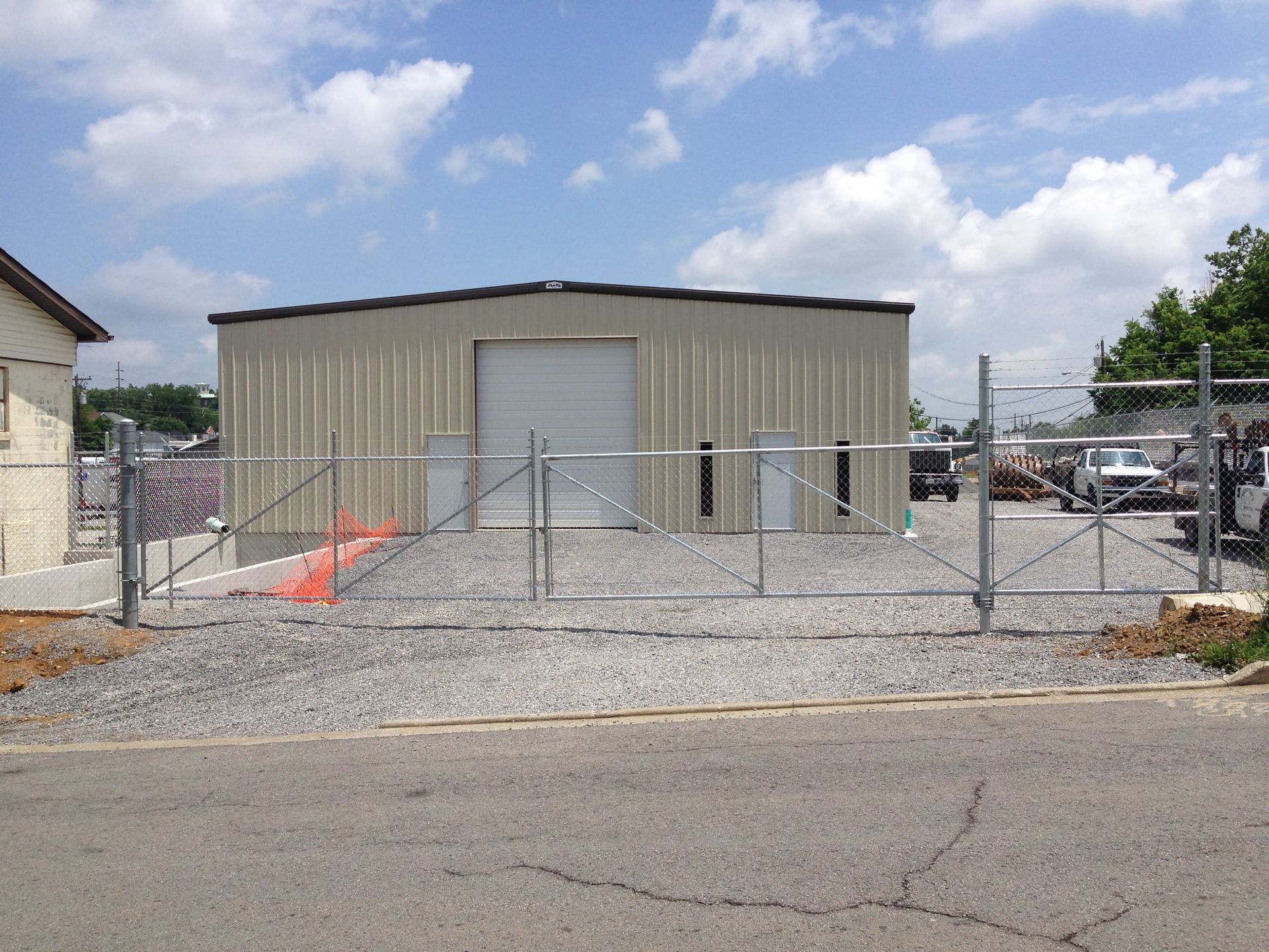 Small tan metal building behind a chain-link fence, with a gravel lot and cars in the background.