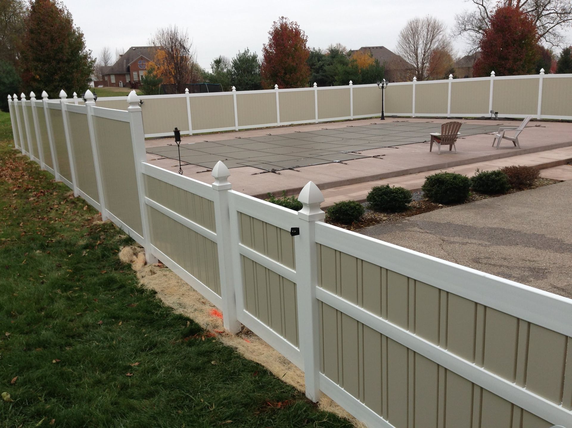 White vinyl fence around a backyard patio with a small dog on a paved area, autumn trees in the background
