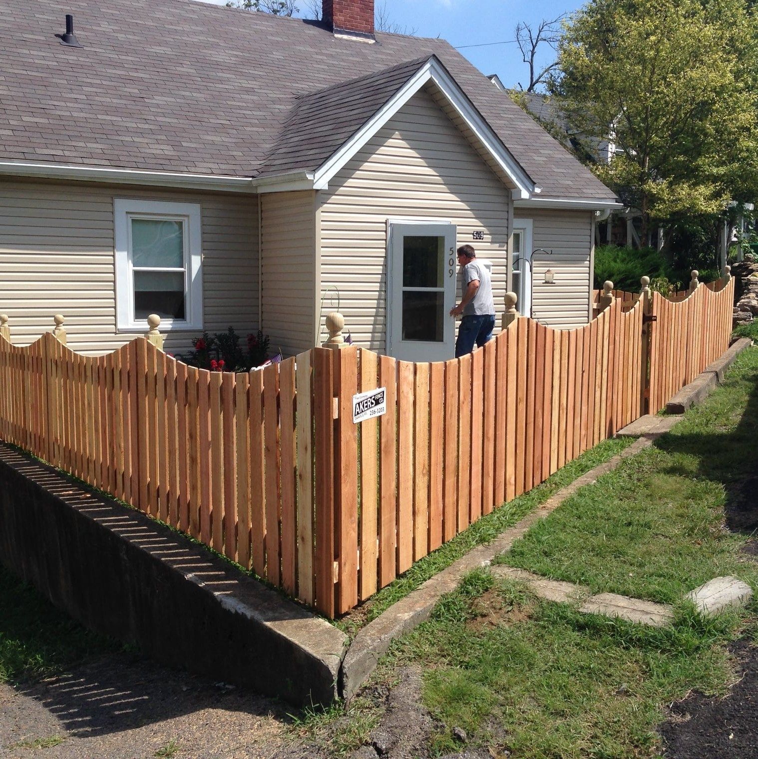 House with new wooden fence; person standing by the side door on a sunny day.
