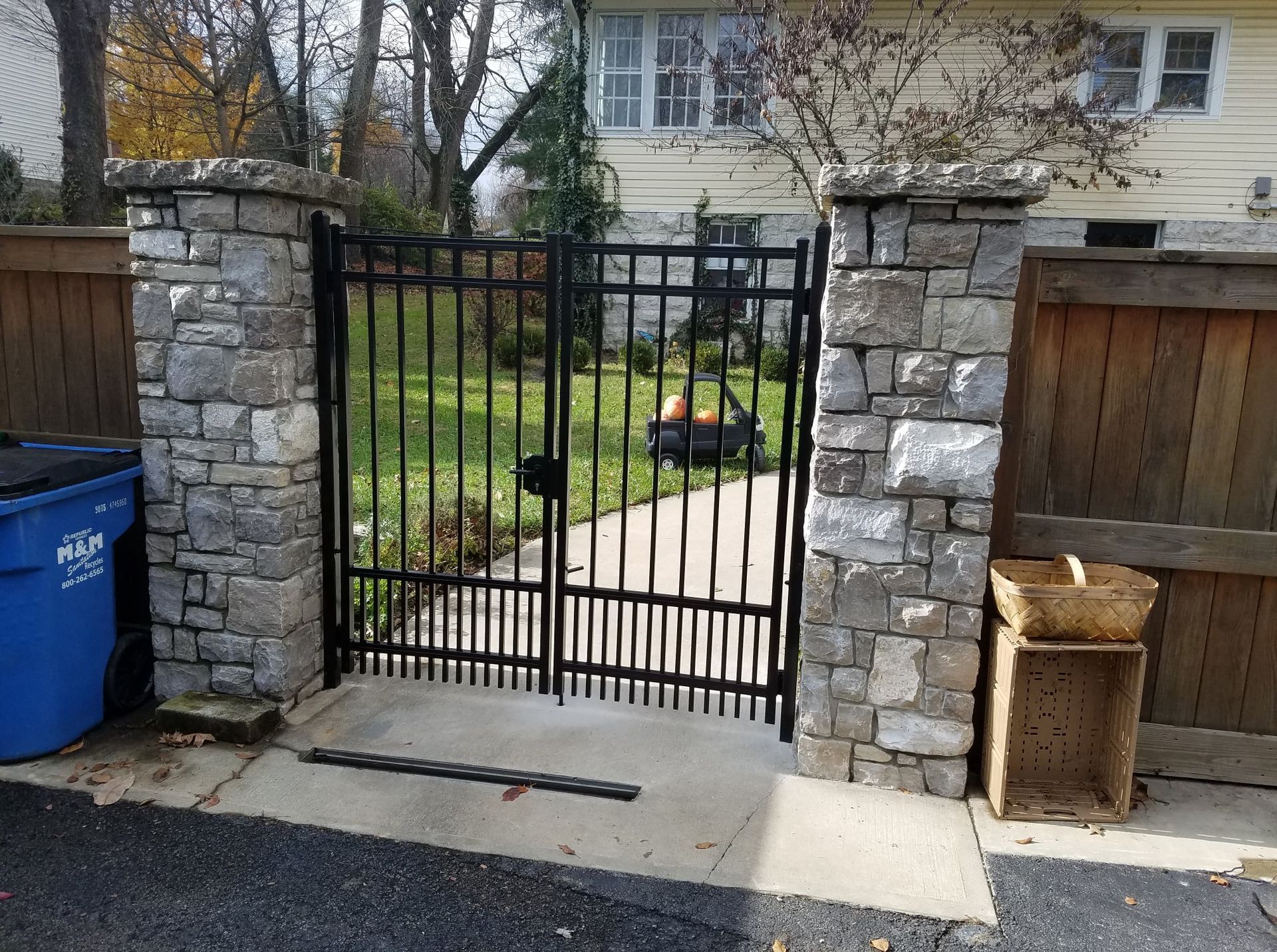 Black metal gate between stone pillars at a driveway entrance, with a blue recycling bin and trash can nearby