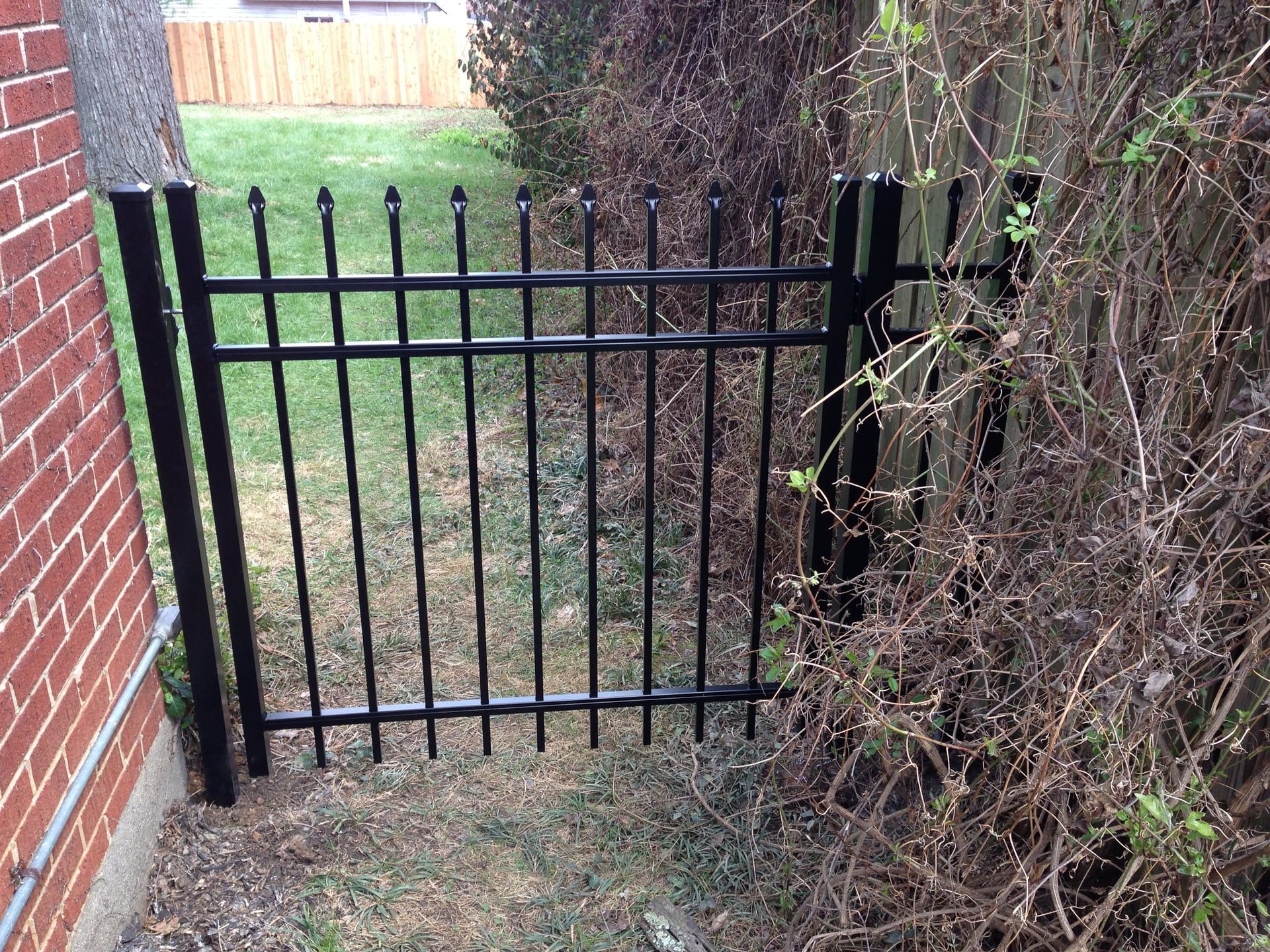 Black metal garden gate between a brick wall and hedge, leading to a yard.