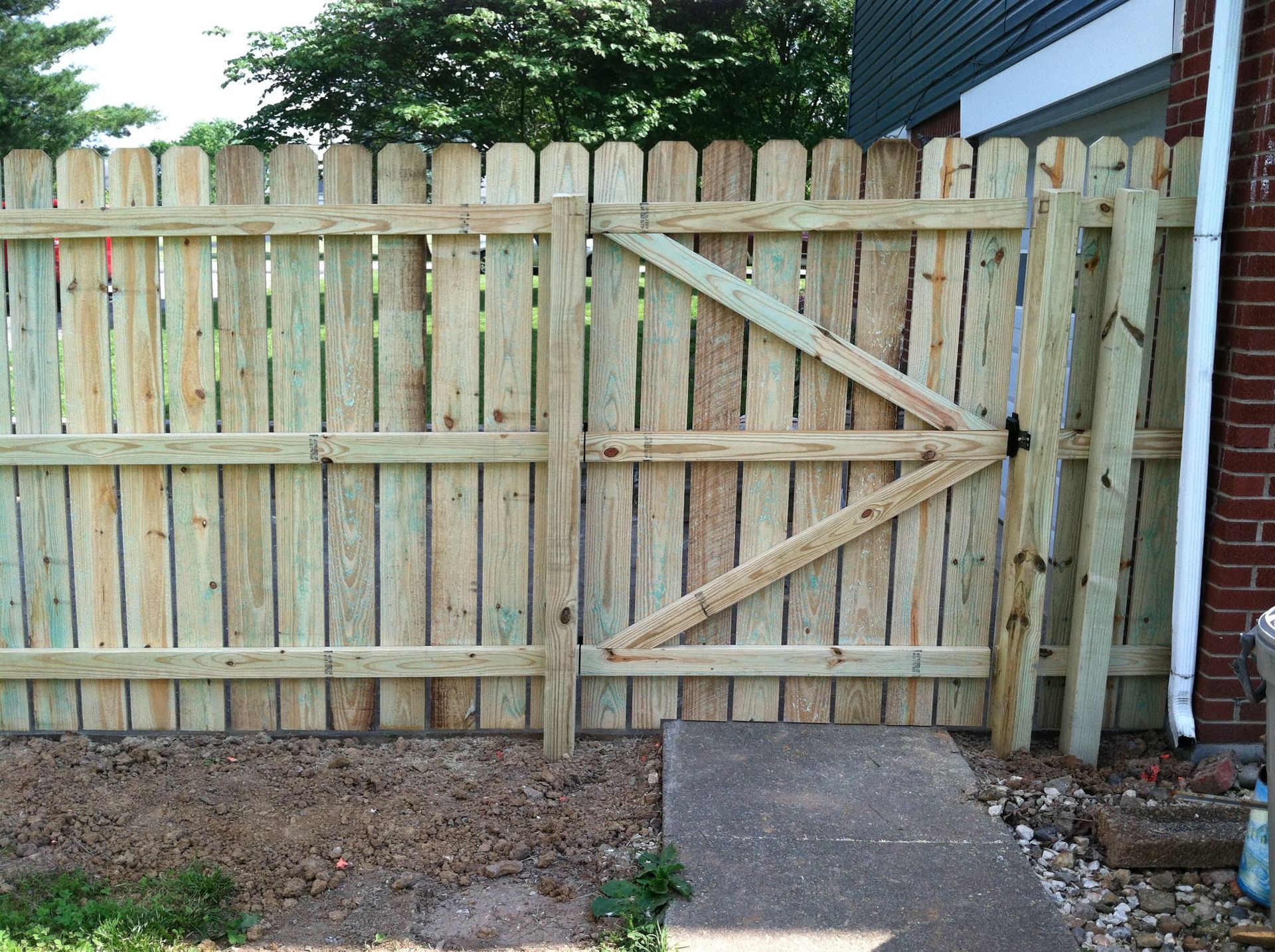 Wooden fence with a diagonal-braced gate beside a brick house and a concrete path