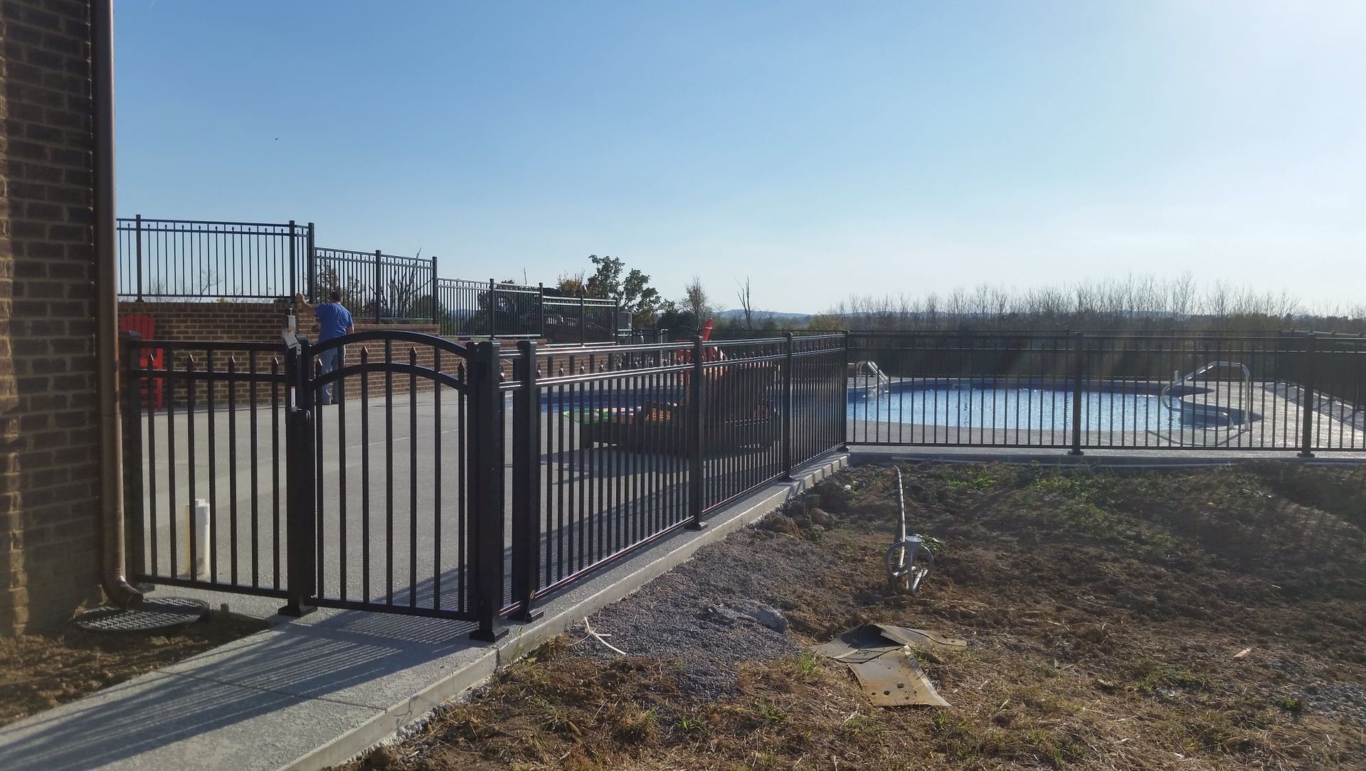 Black metal fence beside a sunny outdoor pool with dry grass and a brick building edge