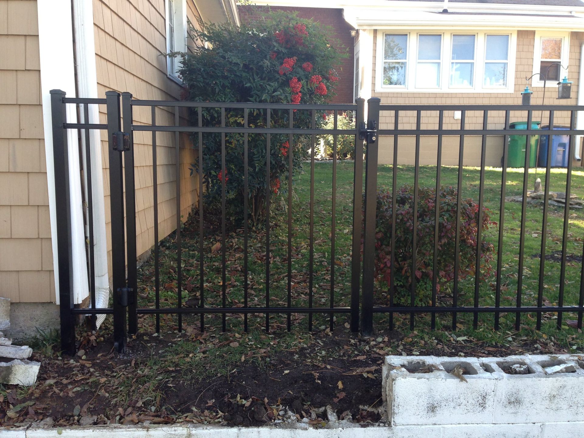 Black metal fence beside a tan house, with shrubs and a small garden bed in front.
