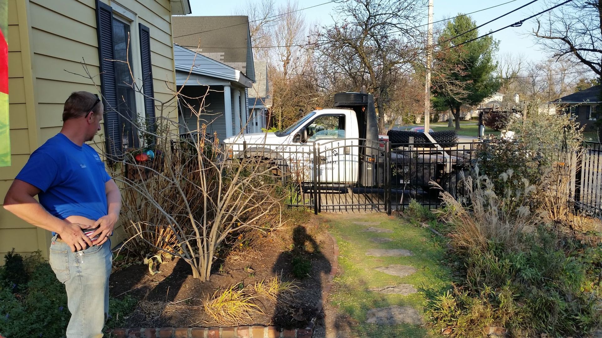 Man standing beside a house, looking toward a small front yard and gate on a sunny day.