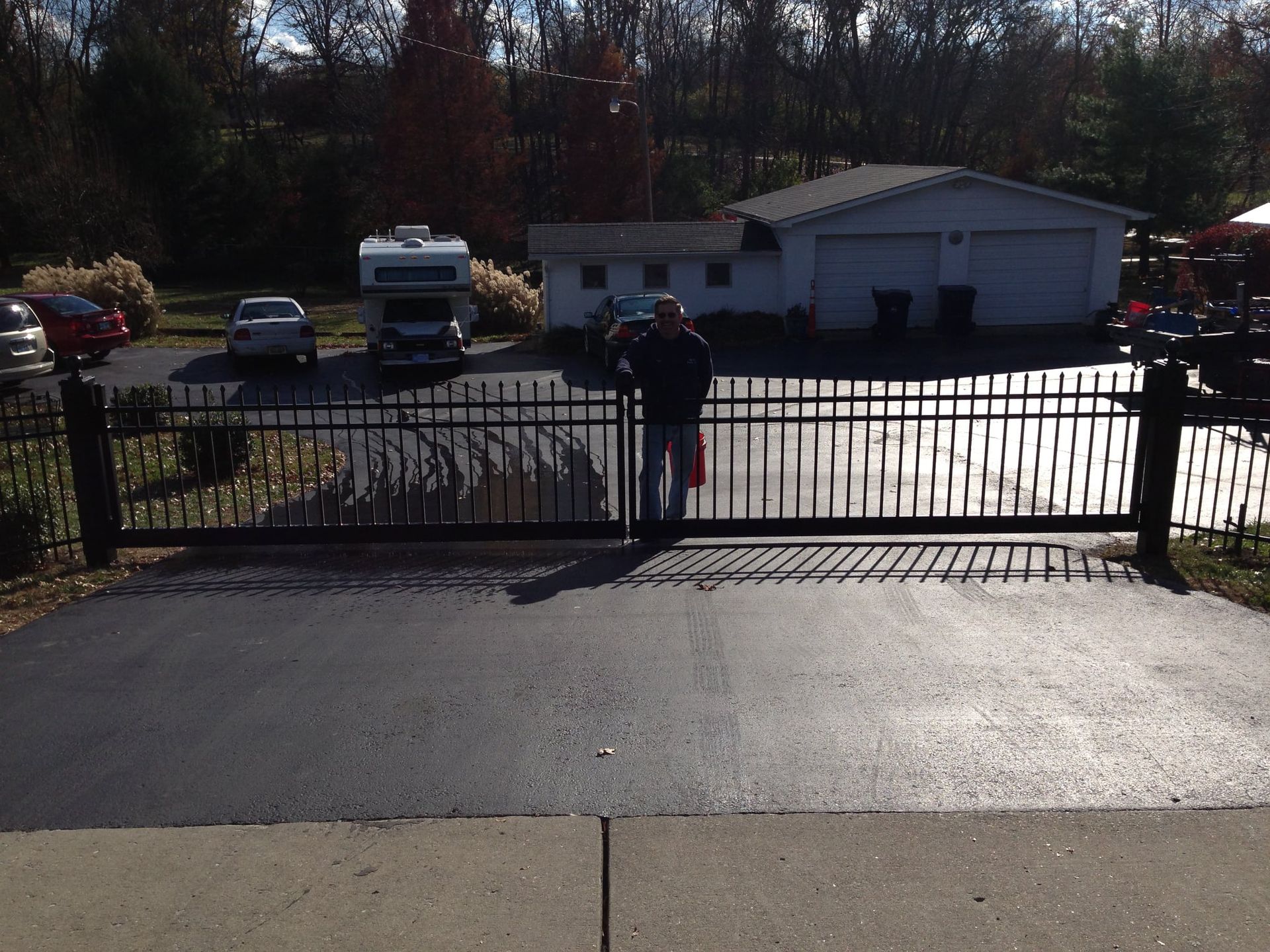 Person standing at a black metal gate in front of a garage and parked cars on a sunny day