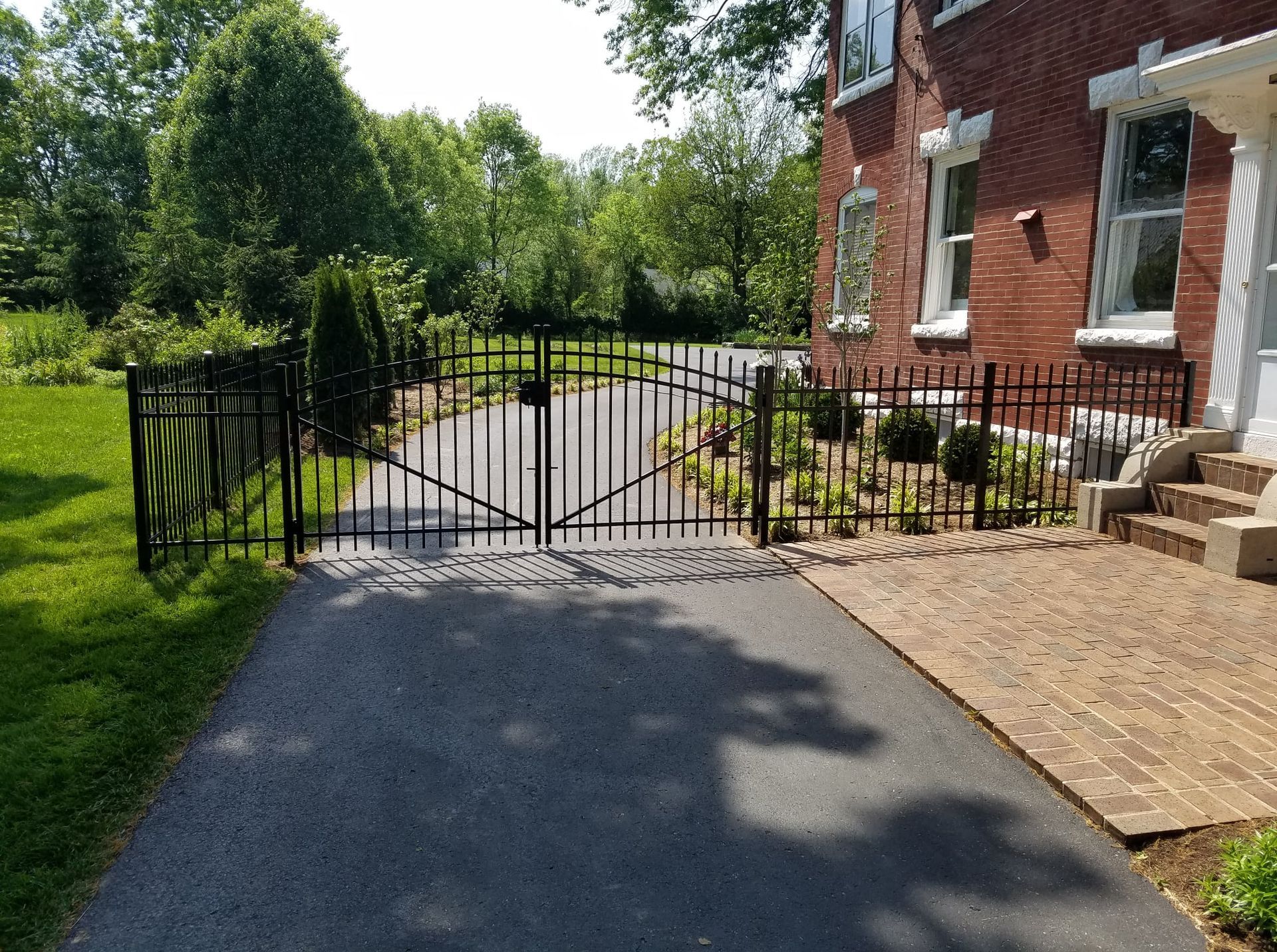 Black metal gate blocking a driveway beside a brick house, with trees and a paved path in the yard.