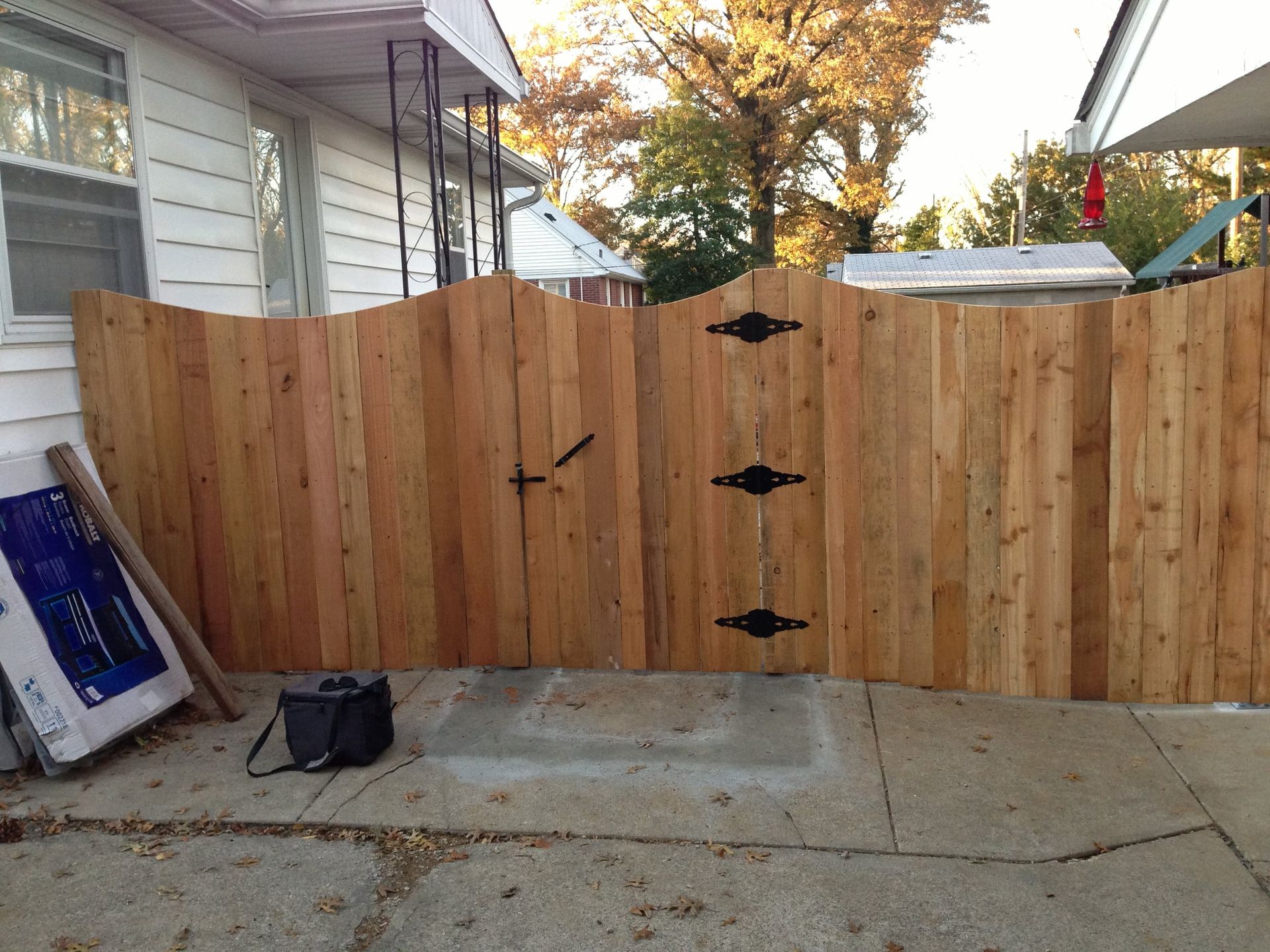 Wooden fence gate across a driveway beside a house, with autumn trees in the background.