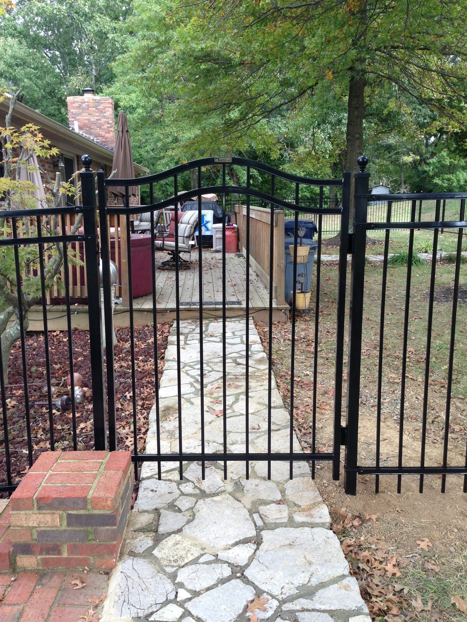 Black metal gate opening to a stone path in a wooded yard, with a red-brick house at left.