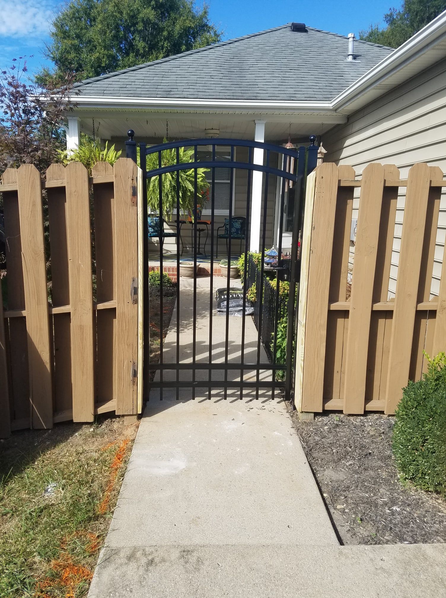Black metal gate in a wooden fence, blocking a concrete walkway to a house driveway.